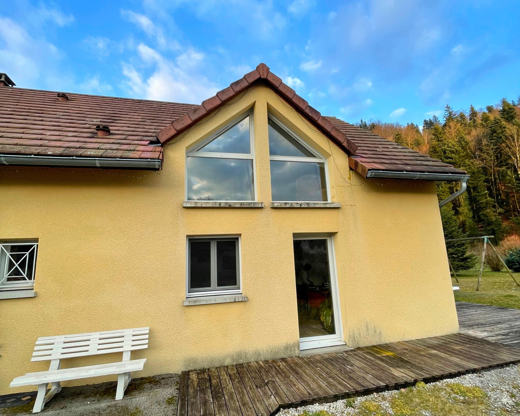 Façade jaune d'une maison avec un toit en tuiles rouges, une porte vitrée, trois fenêtres, un banc blanc et un chemin en bois devant, entourée d'arbres sous un ciel bleu avec des nuages.
