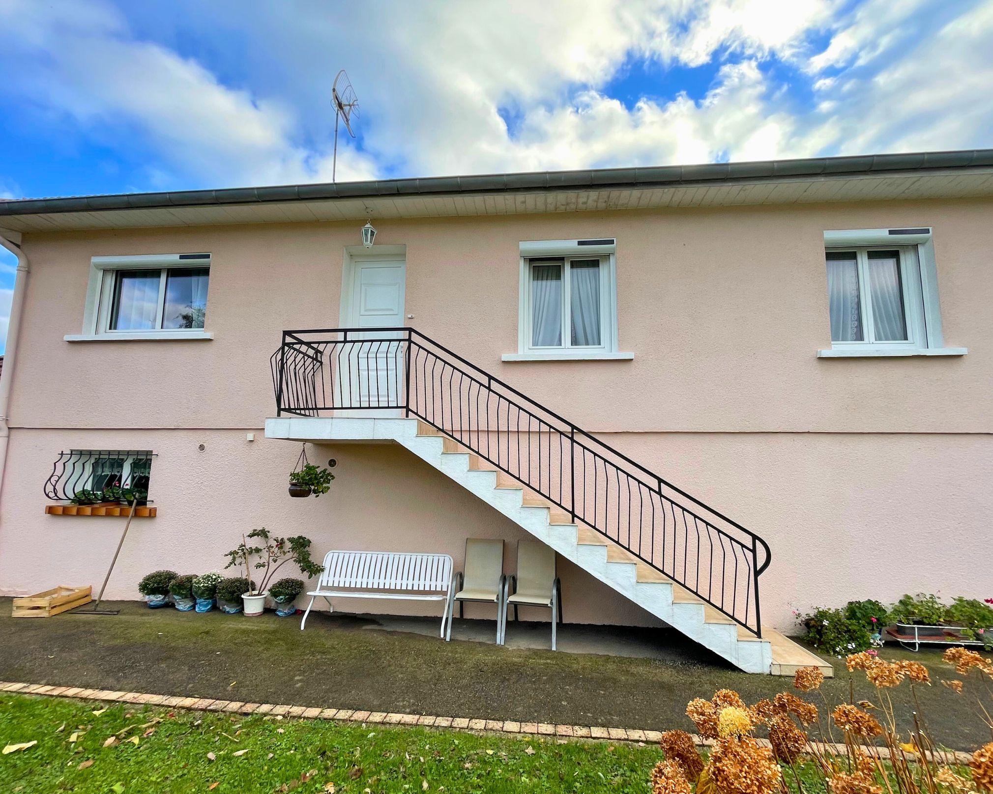 Façade d'une maison de couleur beige avec un escalier extérieur en béton menant à une porte blanche au premier étage, trois fenêtres, des plantes en pots, un banc blanc, deux chaises et un ciel partiellement nuageux bleu.