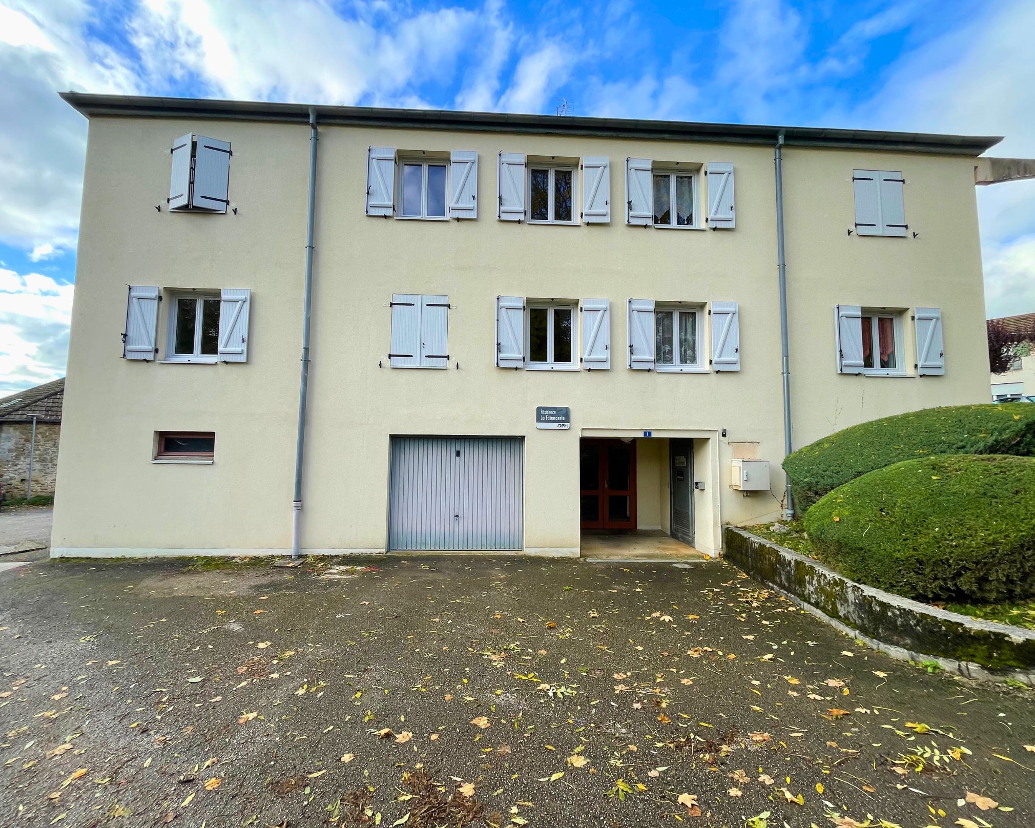 Façade beige d'un immeuble résidentiel avec plusieurs fenêtres équipées de volets blancs, une porte d'entrée, un garage fermé, et des arbustes taillés sur la droite sous un ciel bleu partiellement nuageux.