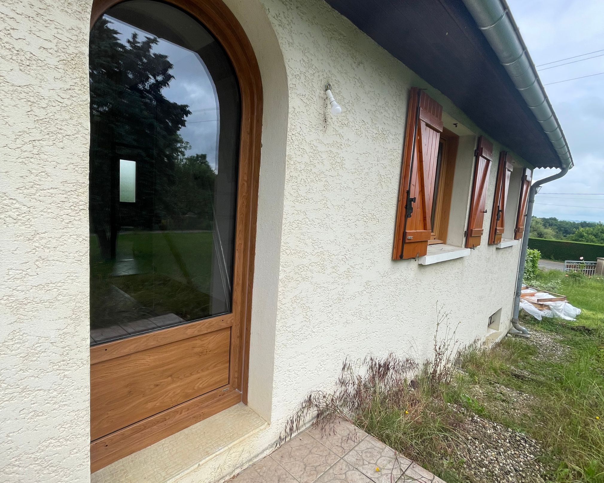 Façade d'une maison avec porte en bois arrondie à gauche, trois fenêtres avec volets en bois ouverts à droite, pelouse et ciel nuageux en arrière-plan.