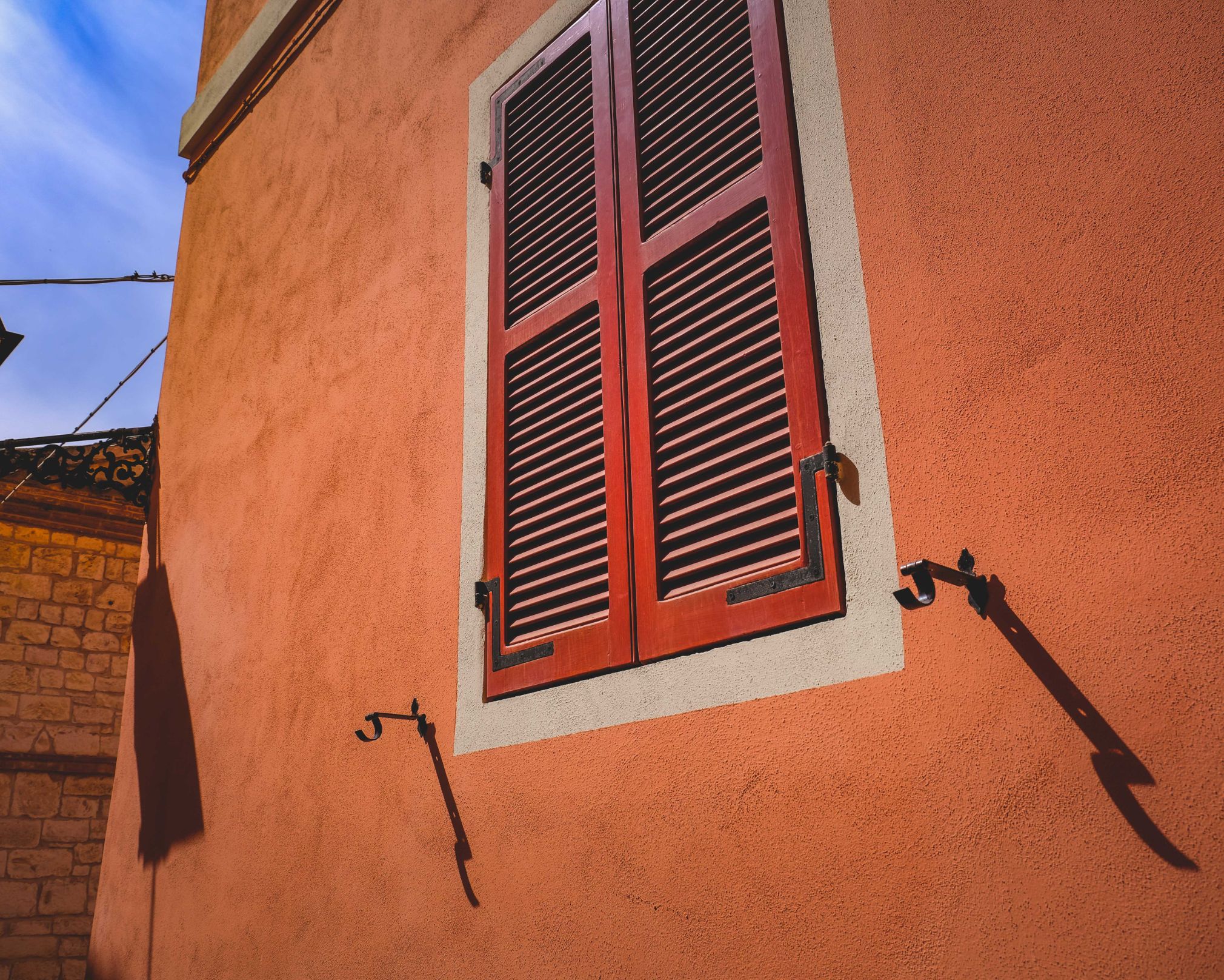 Volet rouge fermé avec des persiennes sur un mur orange texturé, avec deux crochets métalliques noirs sur le mur et un ciel bleu en arrière-plan.