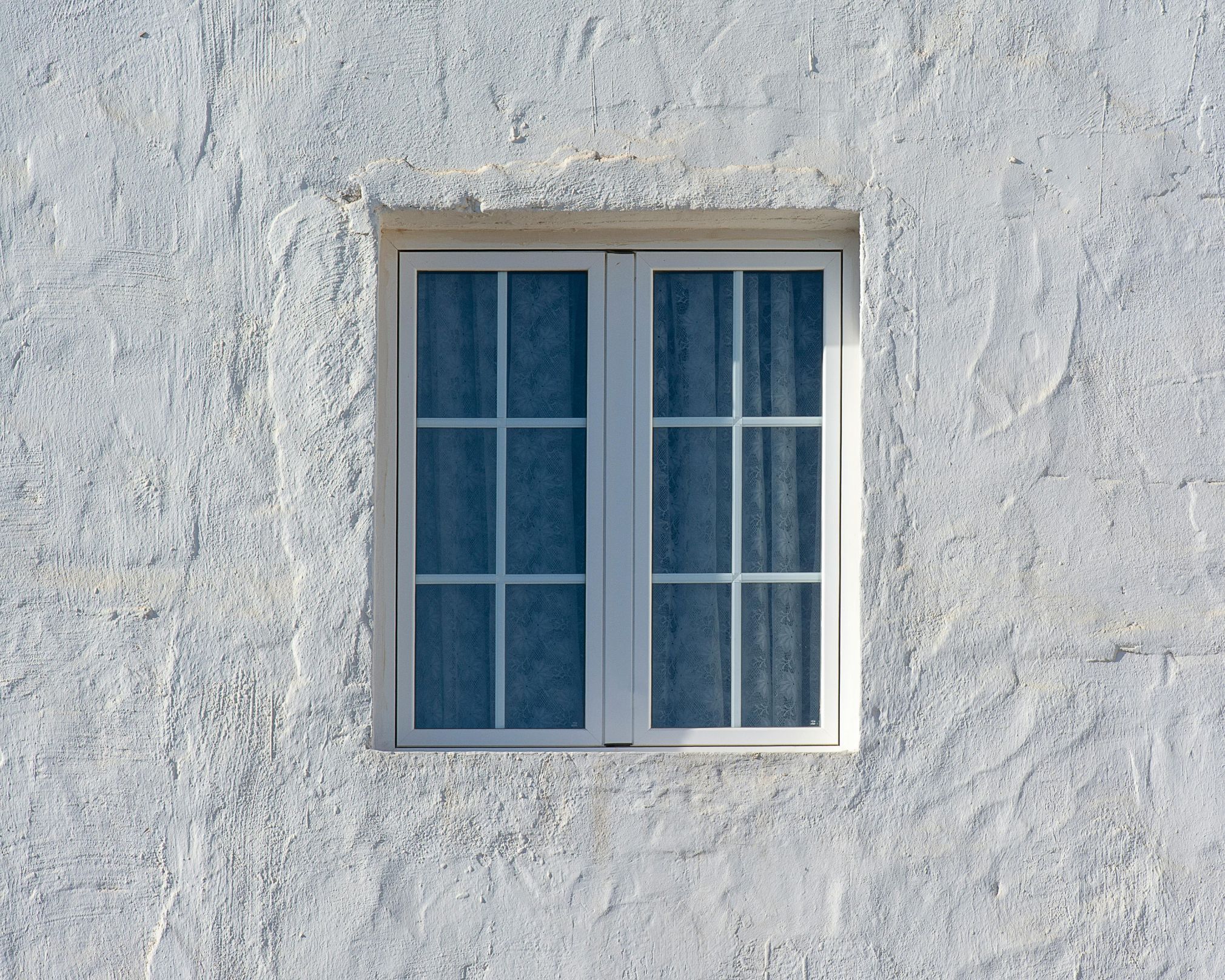 Fenêtre blanche à deux battants avec des rideaux bleus à motifs, encadrée par un mur texturé peint en blanc.