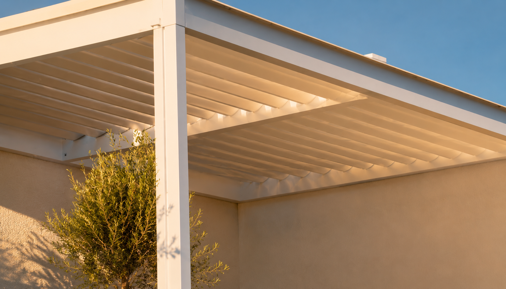 Une pergola moderne blanche avec des lames inclinables, attachée à un mur beige, éclairée par la lumière chaude du soleil avec une plante verte devant.