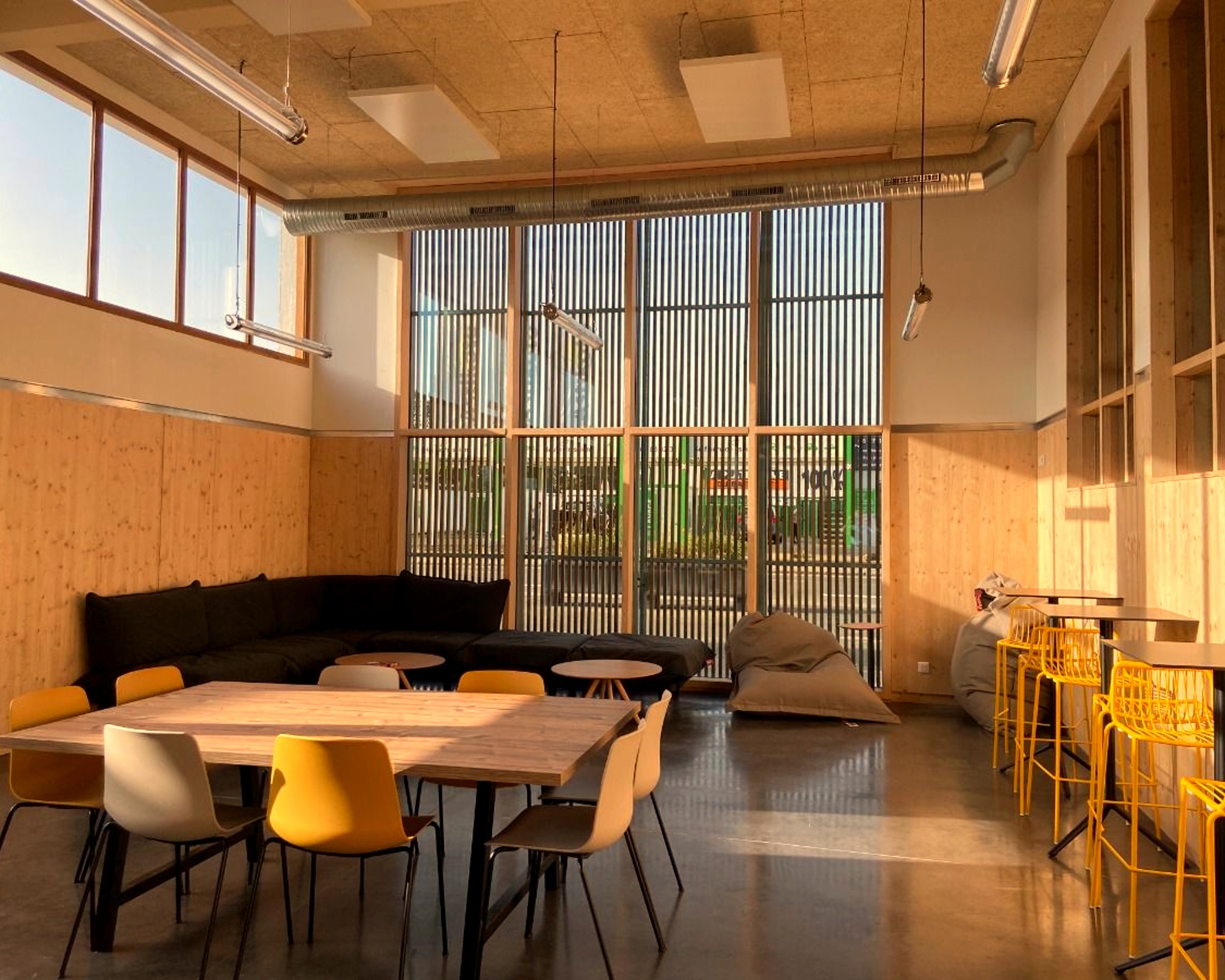 Salle lumineuse avec grandes fenêtres, tables, chaises jaunes et beiges, canapé noir, et poufs gris.