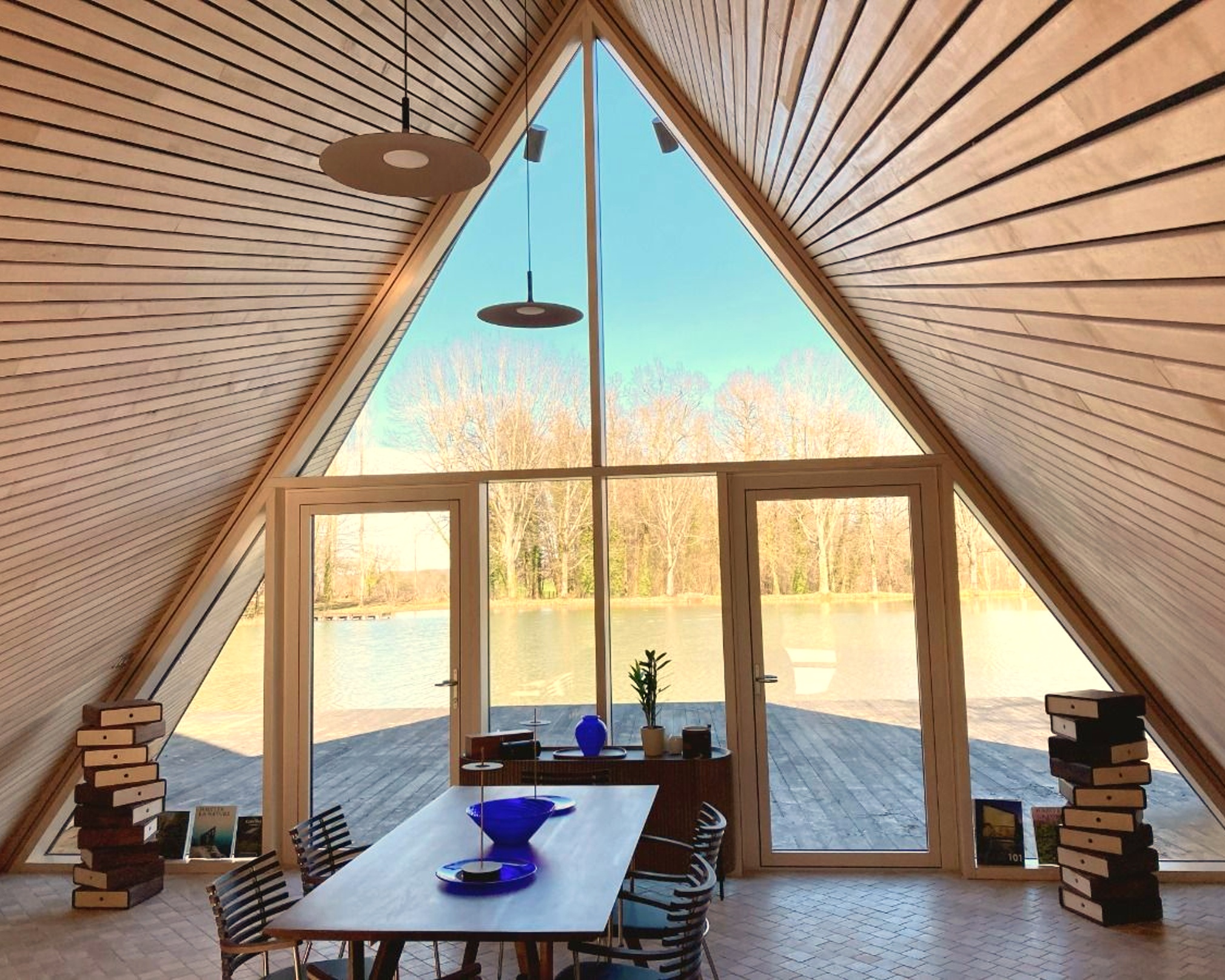 Intérieur d'une maison en forme de A en bois avec table, chaises et grande baie vitrée donnant sur un lac.