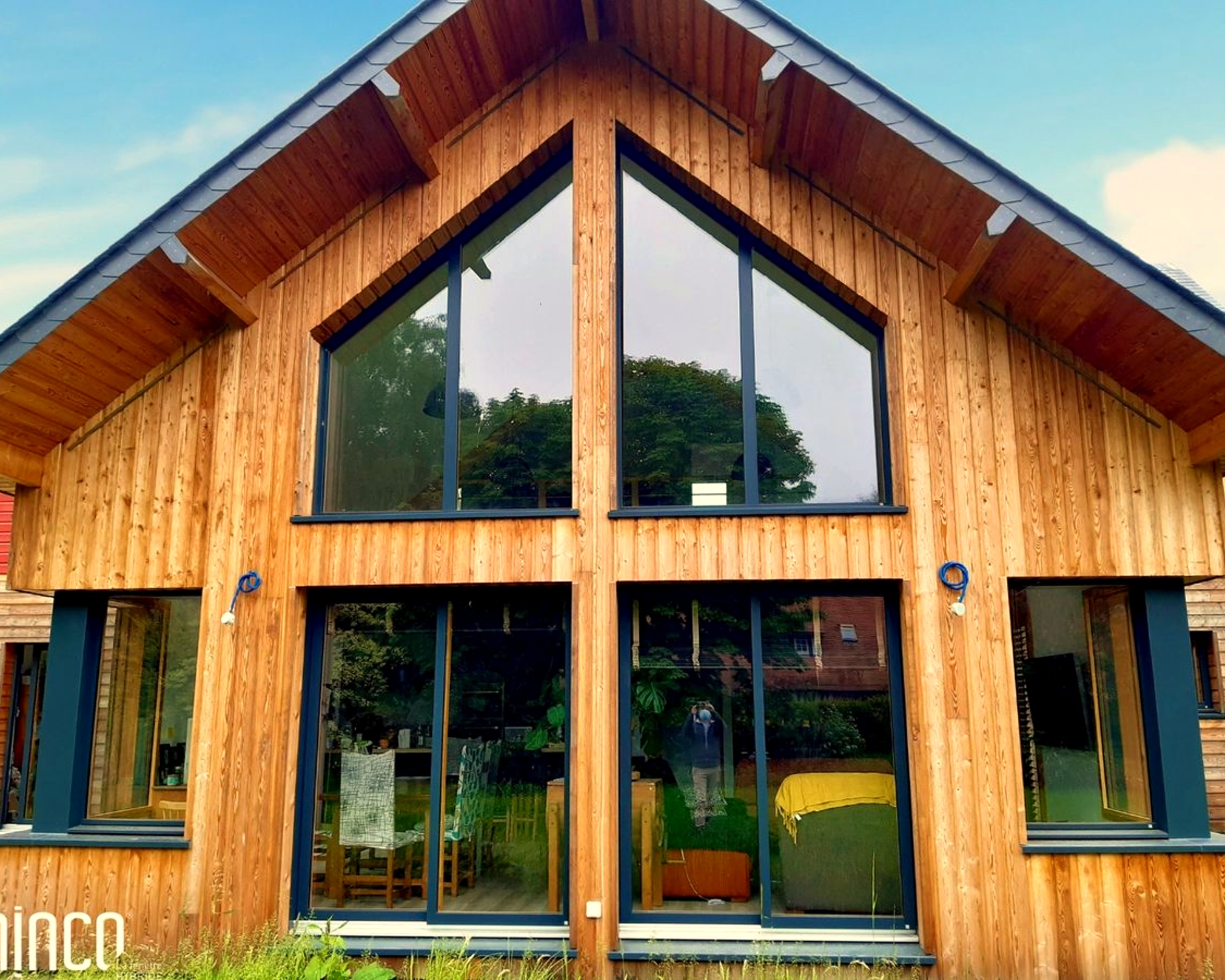 Façade en bois d'une maison avec grandes fenêtres et reflet d'arbres dans le verre.