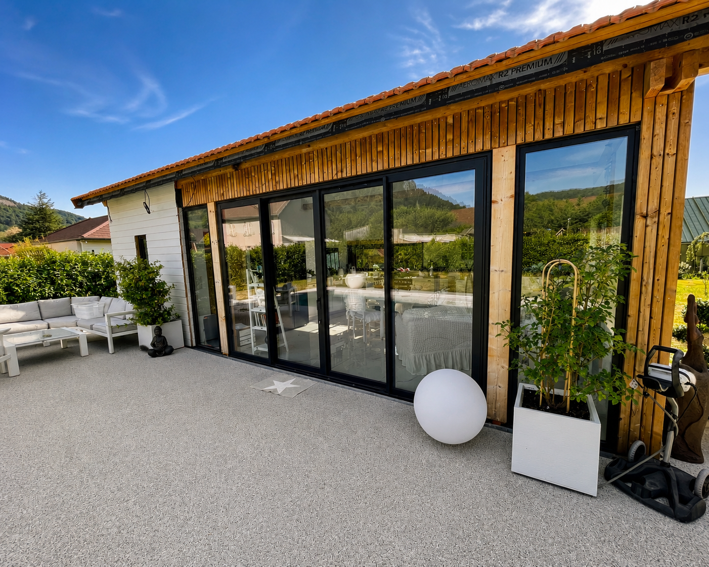 Terrasse ensoleillée avec canapé blanc, grandes fenêtres vitrées d'une maison en bois, plantes en pots et ciel bleu.