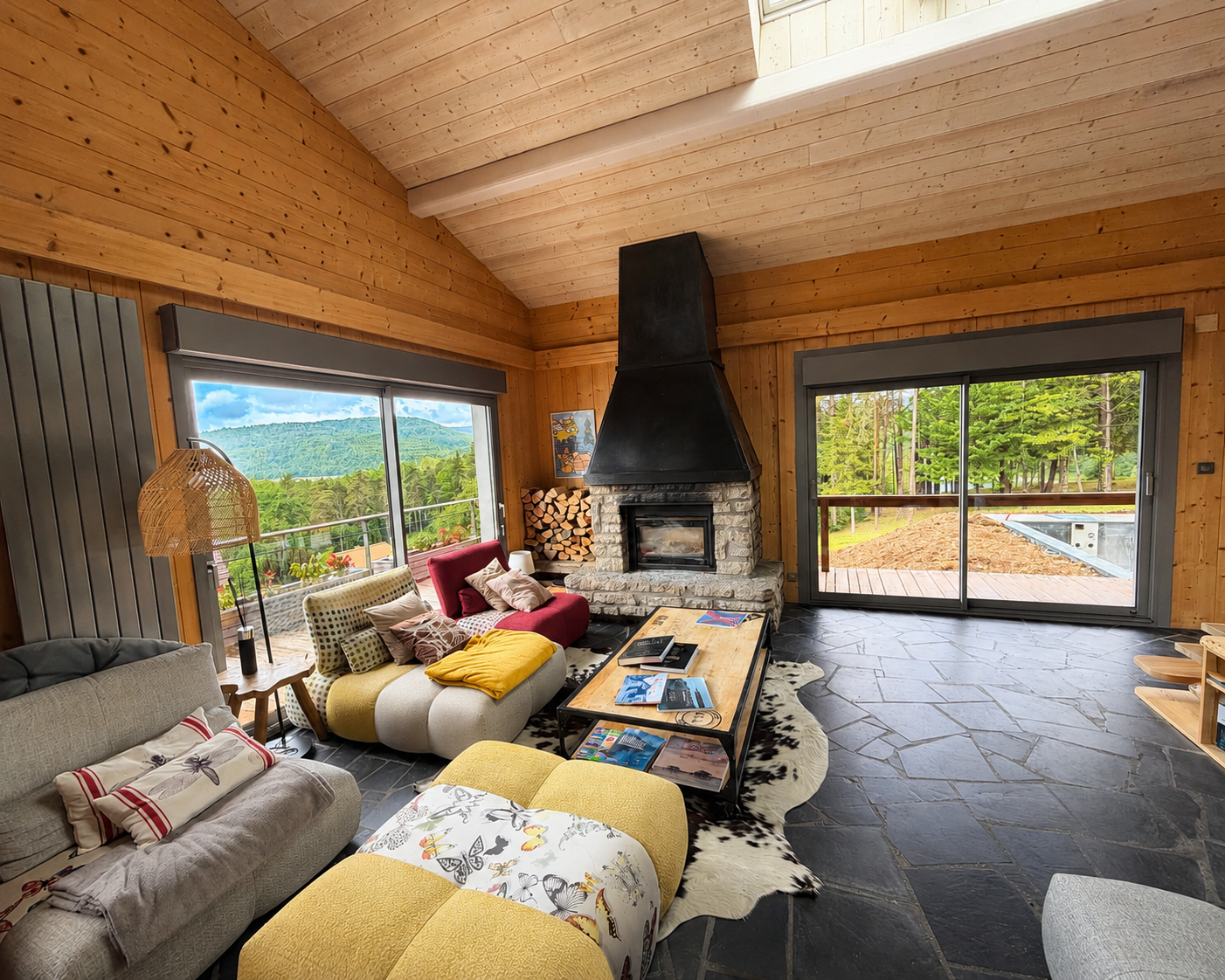 Salon en bois avec un grand foyer central en pierre et métal, canapés colorés avec coussins et couverture, table basse en bois sur tapis peau de vache, grande fenêtre donnant sur une vue montagneuse et forêt verte.