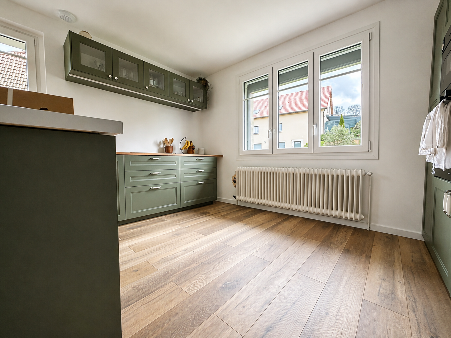 Cuisine moderne avec des armoires vertes, un plan de travail en bois, un radiateur blanc sous une grande fenêtre avec vue sur des maisons voisines, et un sol en parquet clair.