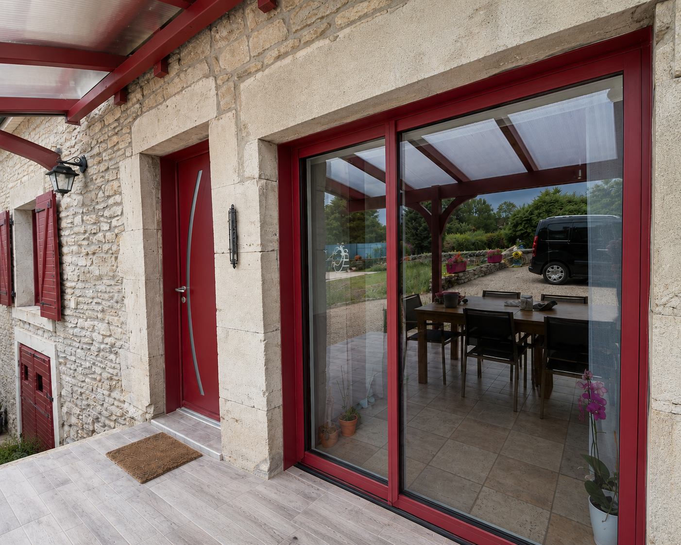 Façade en pierre d'une maison avec porte d'entrée rouge moderne, grandes portes-fenêtres coulissantes rouges donnant sur une terrasse couverte avec table et chaises, plantes en pot et un van noir stationné à l'extérieur.