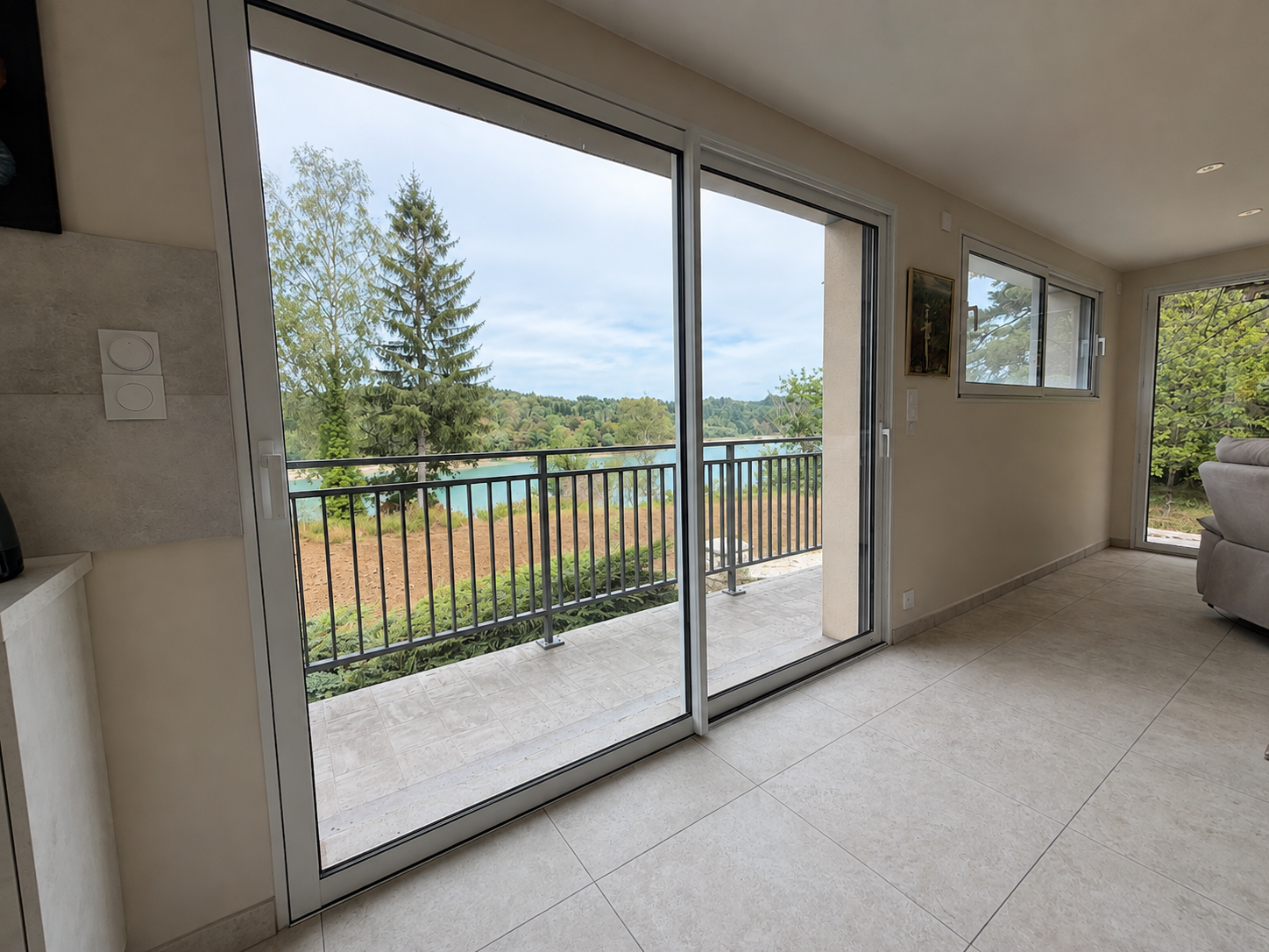 Vue d'un salon avec sol carrelé beige et portes vitrées coulissantes donnant sur un balcon avec balustrade noire et vue sur un lac et des arbres verts.