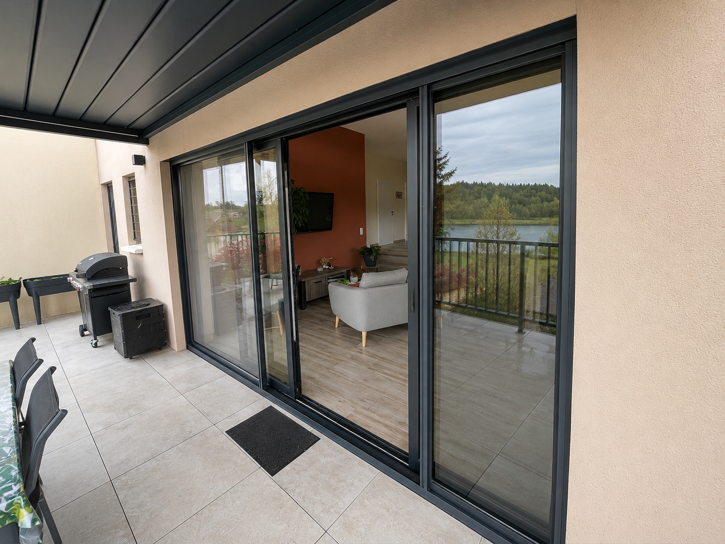Terrasse carrelée avec une table, un barbecue, et des jardinières, donnant sur un salon intérieur visible par une grande baie vitrée coulissante et une vue sur un lac et une forêt.