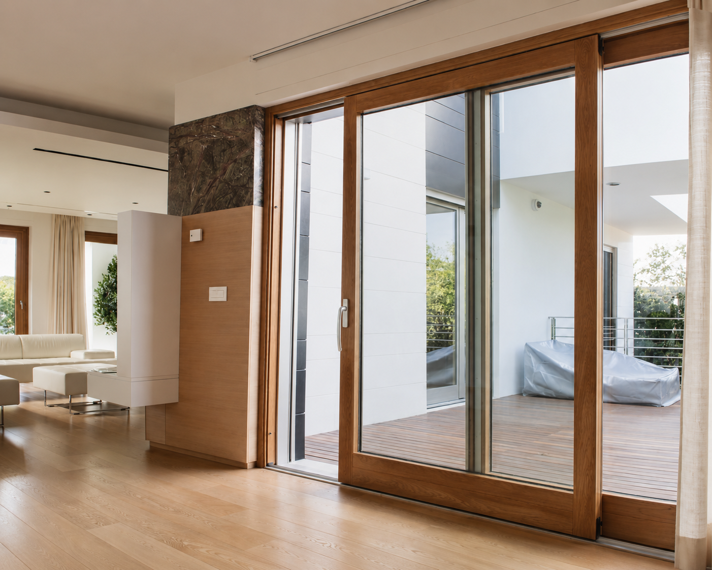 Intérieur lumineux avec sol en bois clair, canapé blanc, rideaux beiges et grande porte-fenêtre coulissante en bois donnant sur une terrasse en bois avec mobilier couvert.