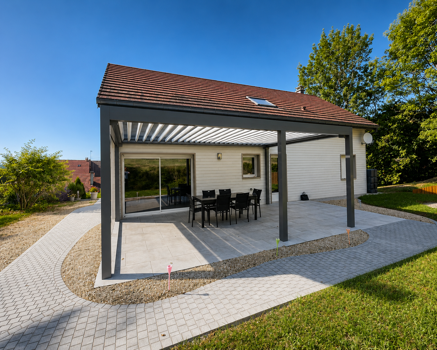 Maison moderne avec une terrasse couverte en métal, une table noire et six chaises, entourée d'un chemin pavé et d'un jardin avec des arbres sous un ciel bleu clair.
