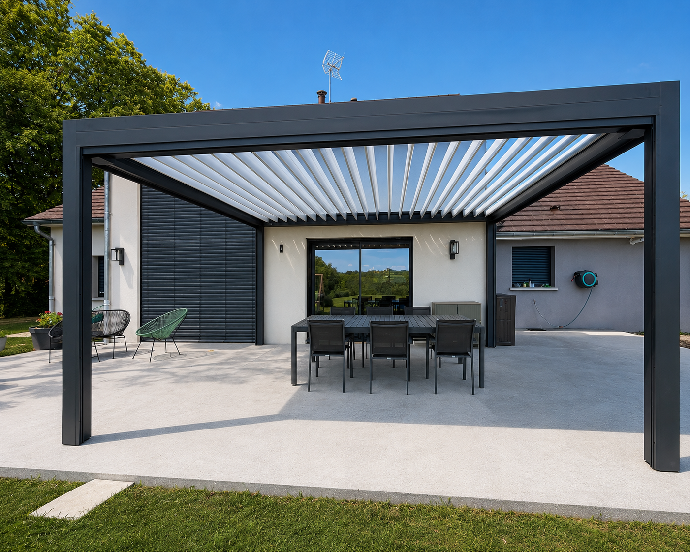 Terrasse de maison moderne avec pergola en métal noir, table à manger noire avec six chaises, deux fauteuils, et grande porte vitrée reflétant un paysage vert sous ciel bleu.