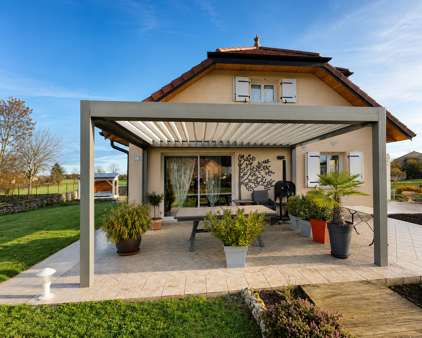 Maison beige avec toiture en tuiles foncées, terrasse carrelée sous pergola moderne en métal, plusieurs grandes plantes en pots, table basse et barbecue, jardin vert et ciel bleu clair en arrière-plan.