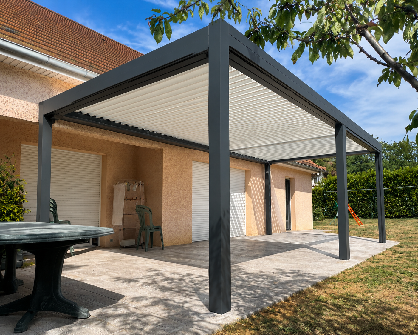 Terrasse avec pergola moderne à lames inclinables en métal gris devant une maison avec volets blancs, table ronde et chaises en plastique vertes, pelouse et ciel bleu.