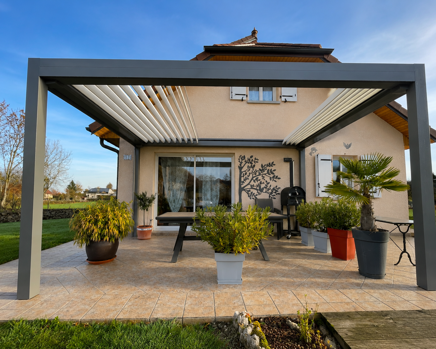 Maison beige avec terrasse carrelée couverte d'une pergola moderne en métal gris, plusieurs pots de plantes, table et barbecue noir, sous un ciel bleu clair.