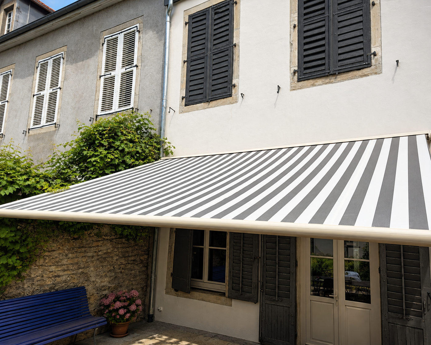 Façade de maison avec fenêtres à volets noirs et blancs. Au premier plan, un auvent rayé noir et blanc couvre une porte en verre. À gauche, un banc bleu et une plante fleurie sont posés devant un mur en pierre partiellement recouvert de végétation verte.