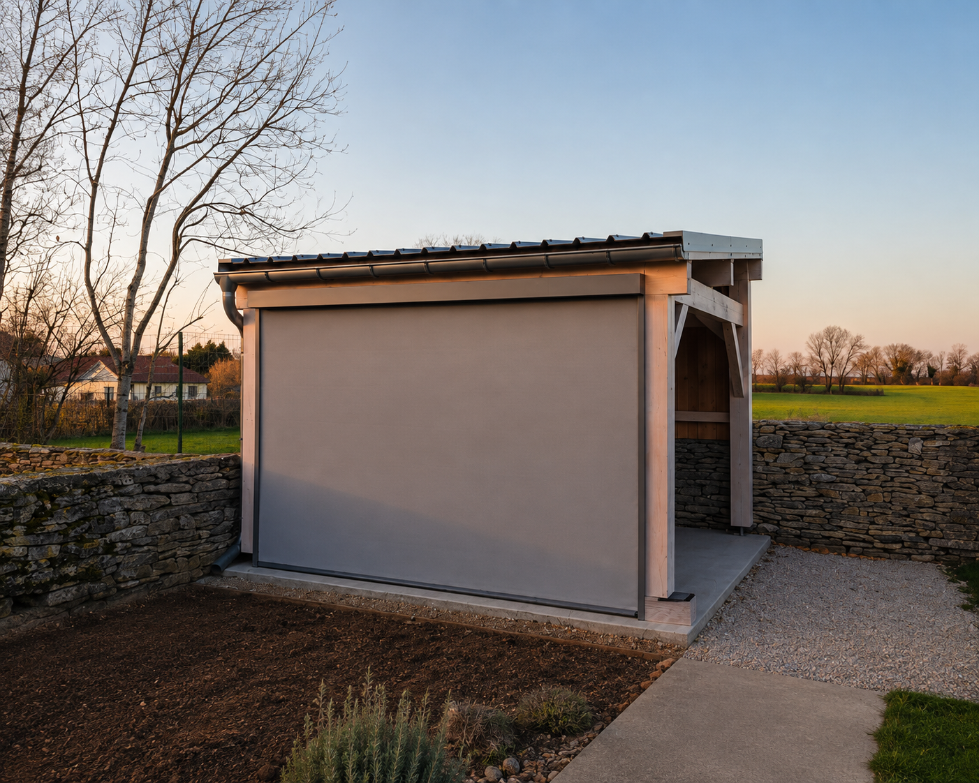 Petit abri en bois avec un auvent et un écran gris tiré, entouré d'un mur en pierre sèche, au coucher du soleil dans un jardin avec des arbres et un champ en arrière-plan.