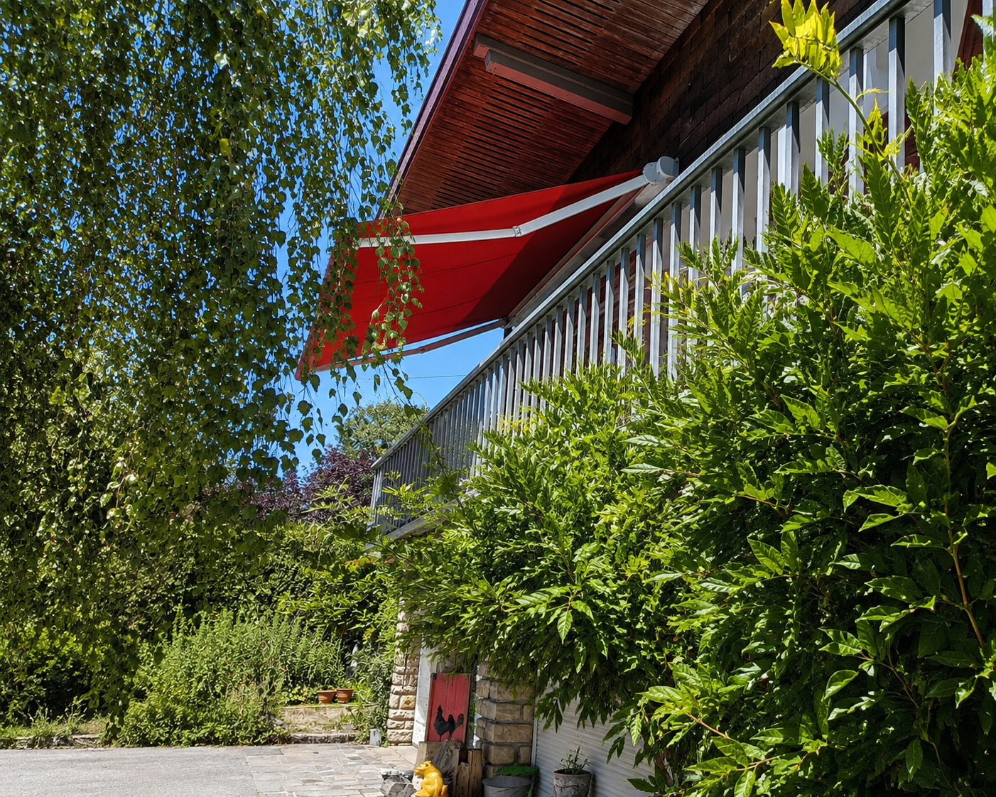 Terrasse d'une maison partiellement visible avec un store rouge déployé, entourée de feuillages verts denses sous un ciel bleu clair.