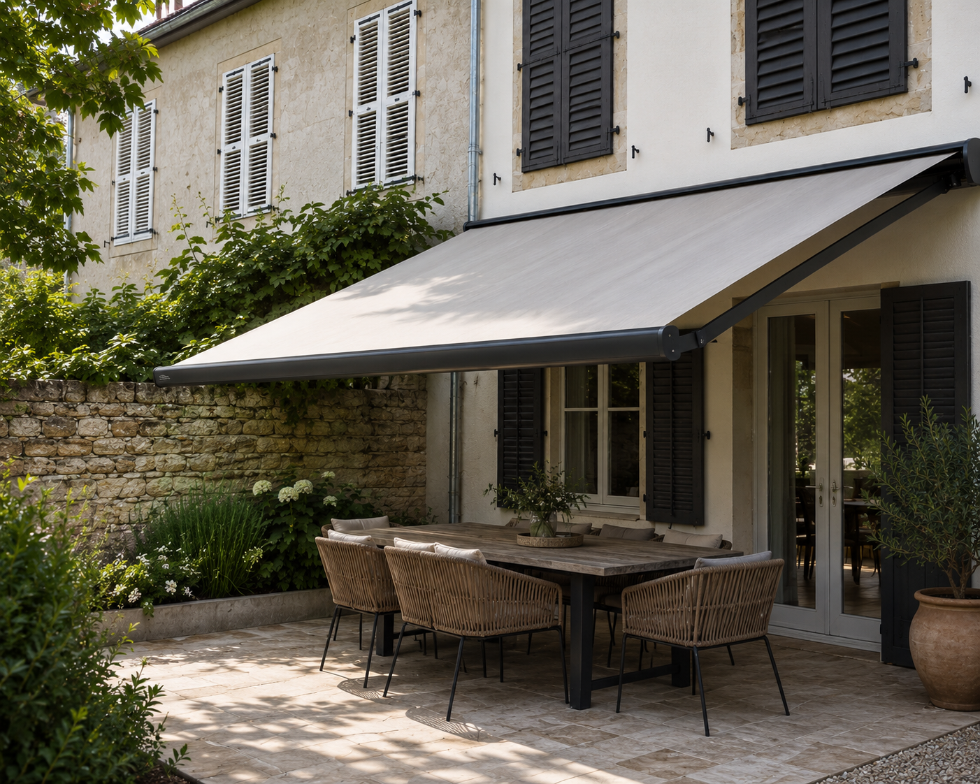 Terrasse extérieure avec table en bois et six chaises en osier sous un store beige fixé à la façade d'une maison avec fenêtres à volets noirs et mur en pierre.