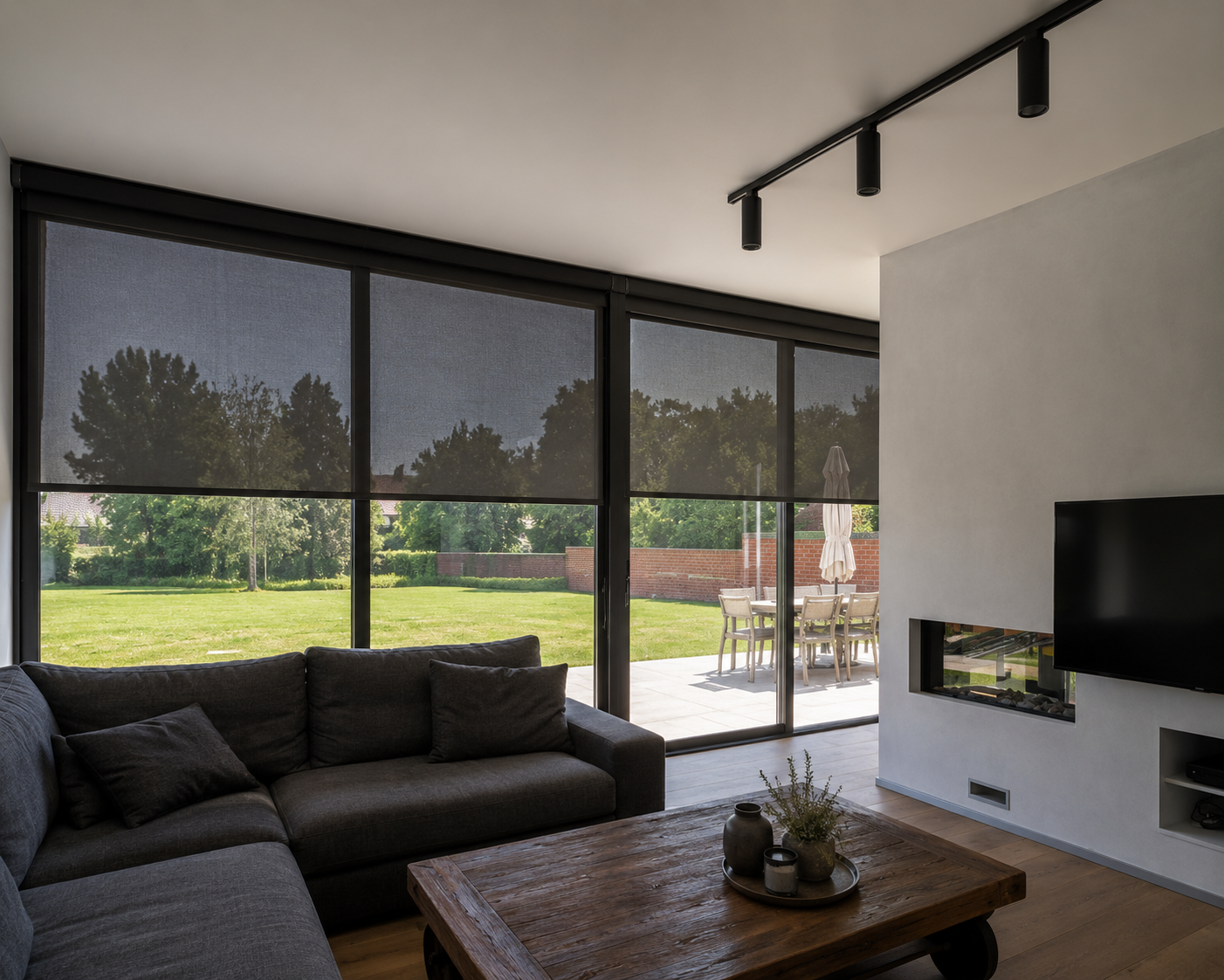 Salon moderne avec canapé gris, grande table basse en bois, cheminée intégrée au mur, télévision murale et grandes fenêtres donnant sur un jardin avec terrasse et mobilier extérieur.