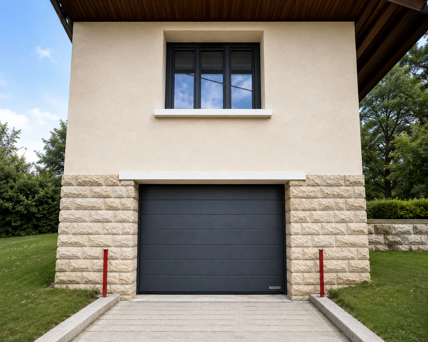 Façade d'une maison avec un garage fermé à porte noire et une fenêtre à trois battants au-dessus, entourée de murs en pierre beige et d'un jardin avec pelouse verte et arbres.