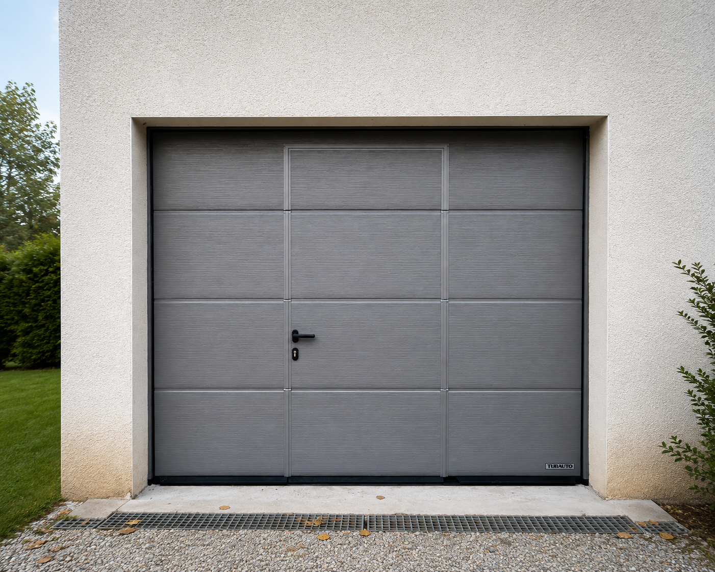 Porte de garage grise moderne avec poignée noire intégrée, encadrée par un mur beige, entourée de pelouse verte et de buissons.
