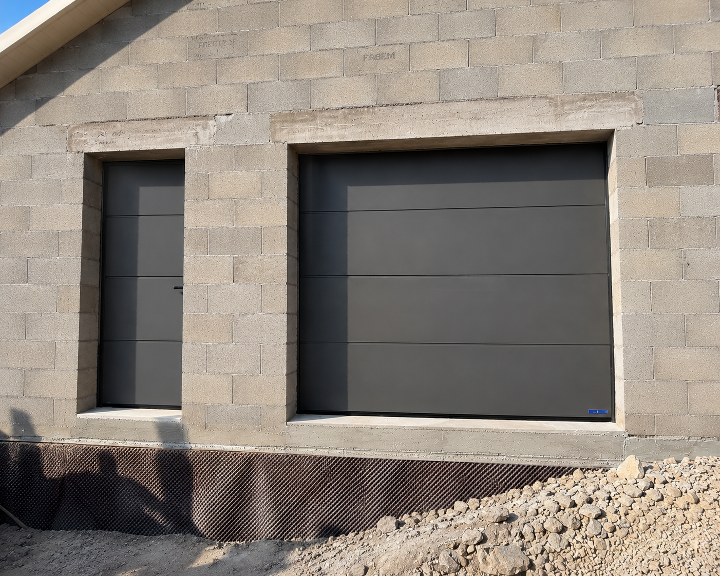 Façade en parpaings avec une porte et une grande fenêtre modernes grises encadrées par le mur en béton, terrain en gravier devant.