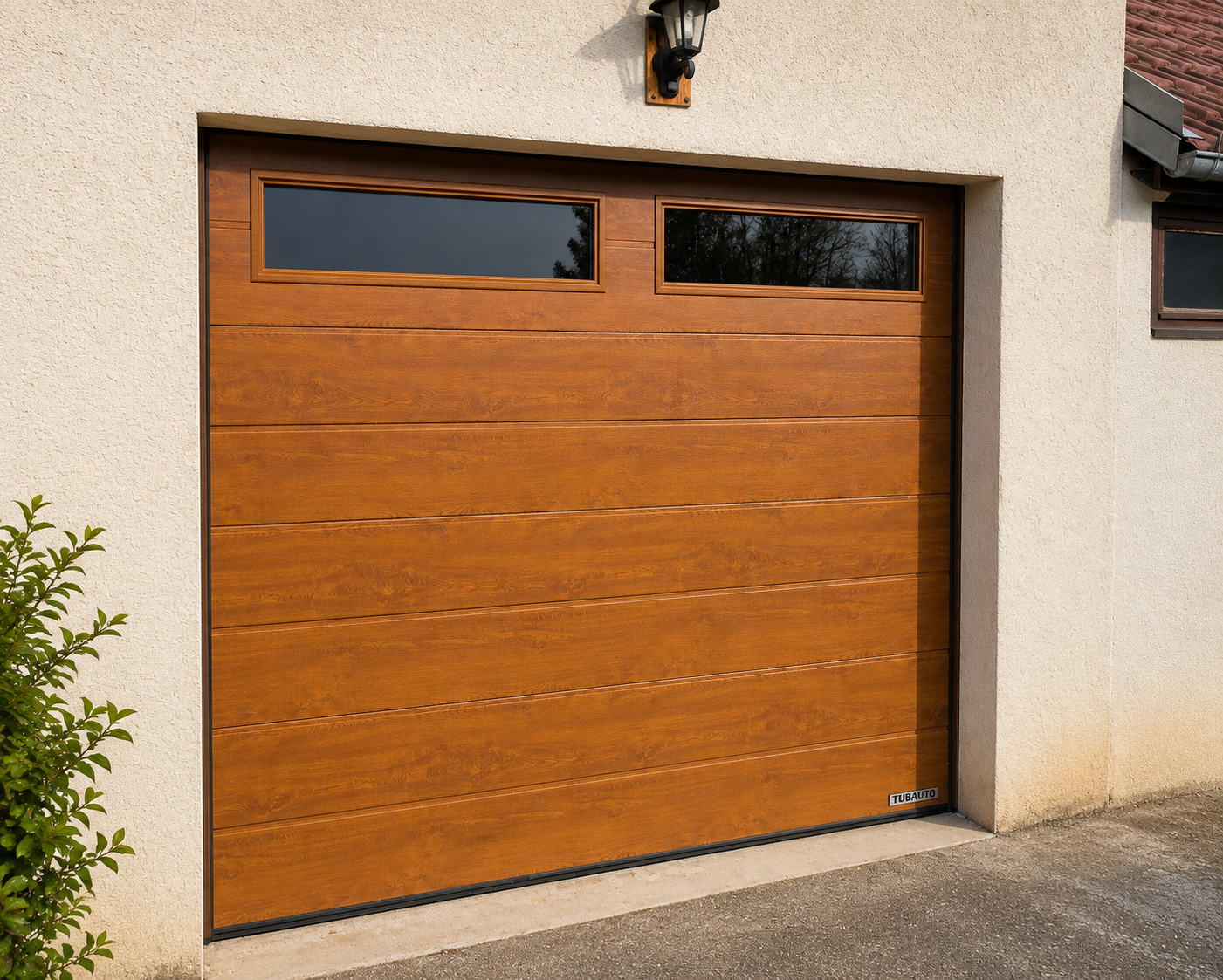 Porte de garage en bois clair avec deux fenêtres rectangulaires en haut, encadrée dans un mur beige avec un petit buisson sur le côté gauche et une lampe noire au-dessus.