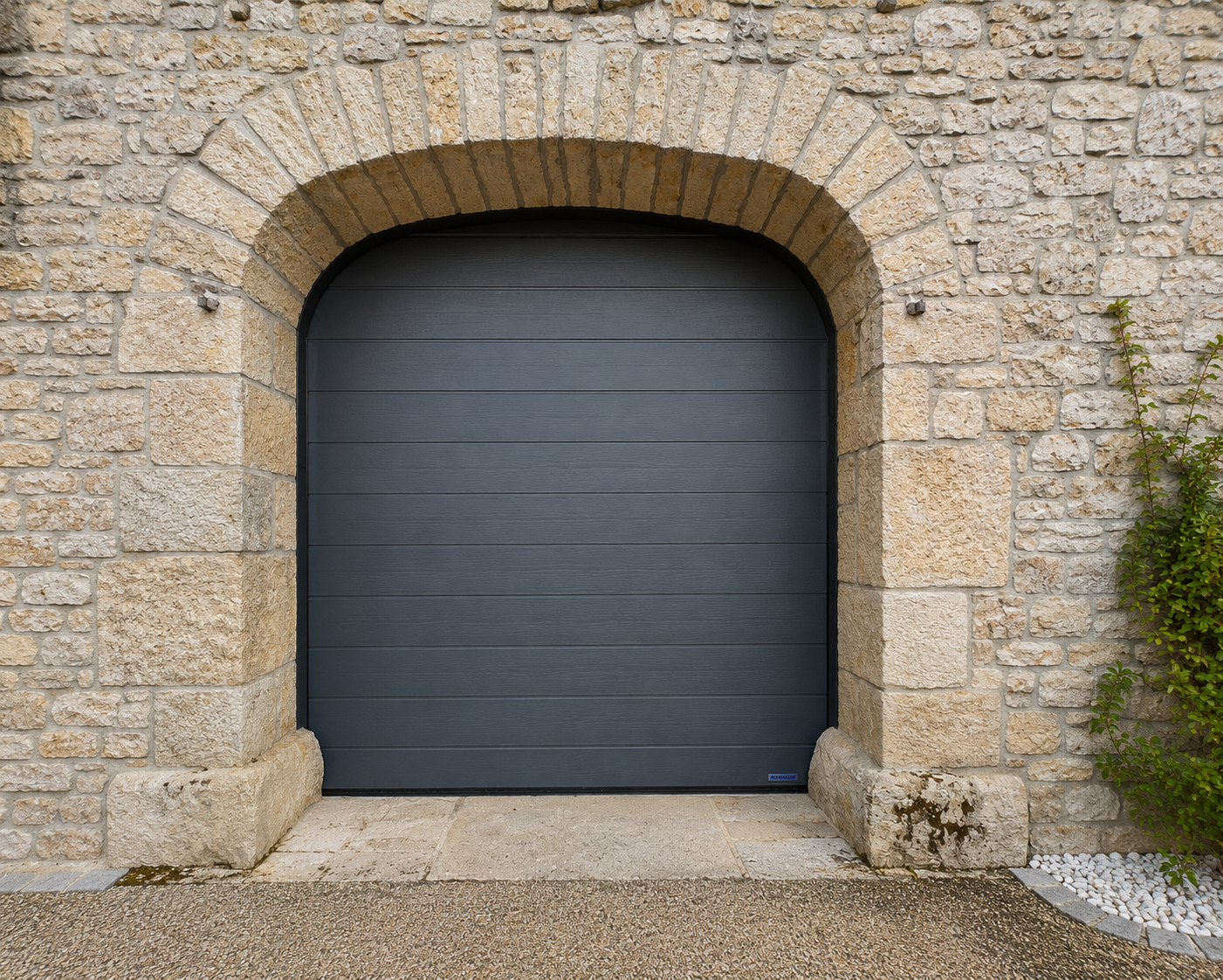 Porte de garage grise avec un arc en pierre dans un mur en pierres claires, bordée de petites pierres blanches et de plantes vertes.
