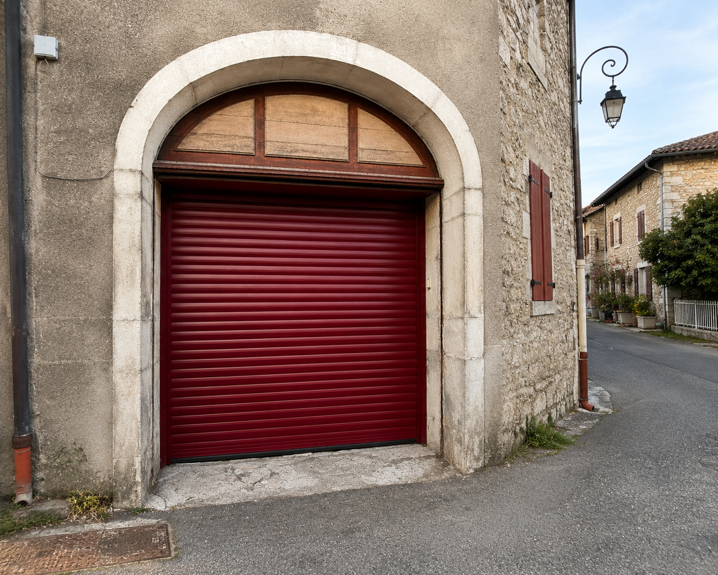Porte de garage rouge foncé en métal avec volet roulant dans un mur en pierre beige ayant une arche blanche, rue pavée calme avec lampadaire et maisons en pierre avec volets rouges.