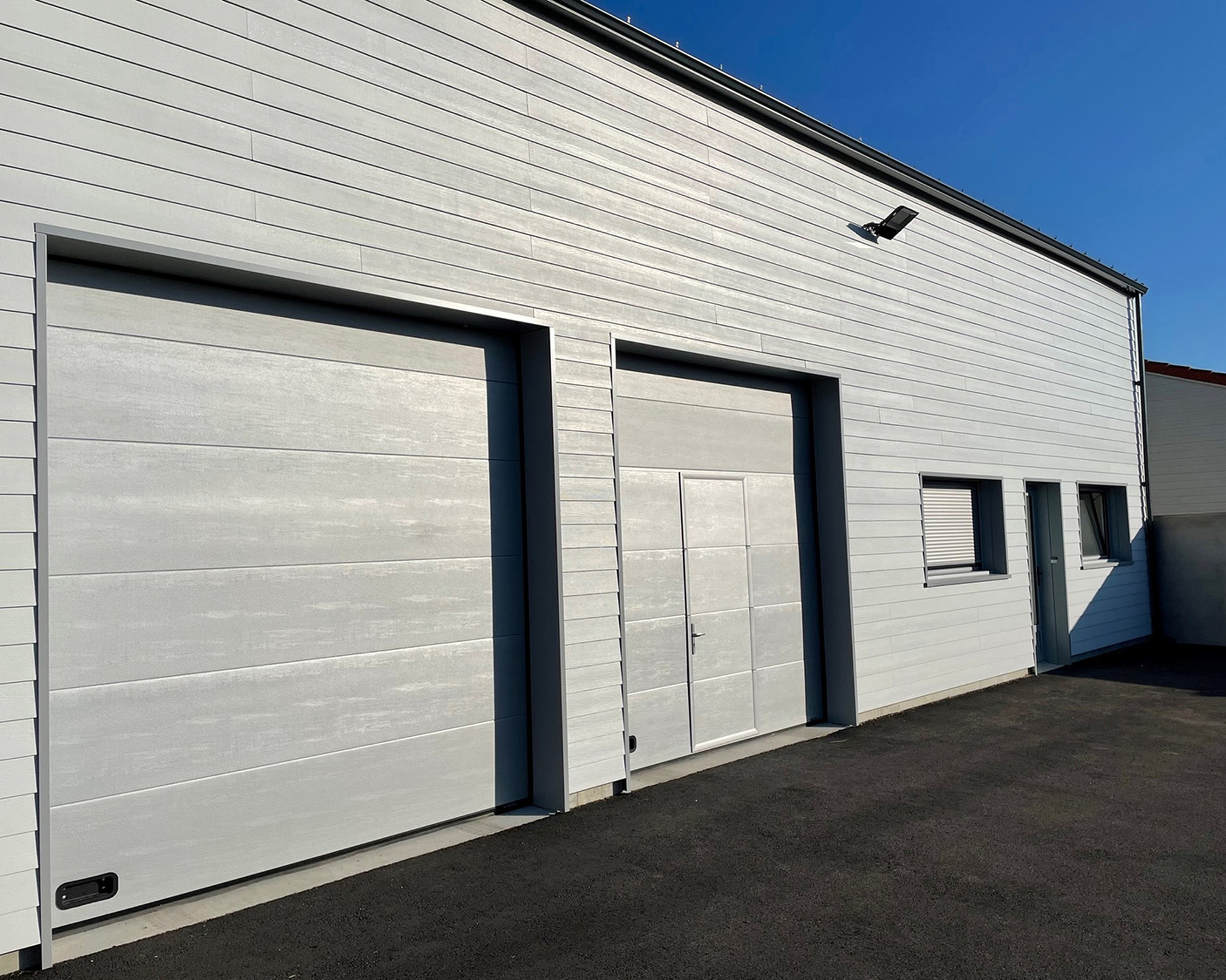 Façade d'un bâtiment industriel gris avec deux grands portails roulants, une porte piétonne, deux fenêtres avec volets fermés et une autre porte grise sur un sol en asphalte noir.