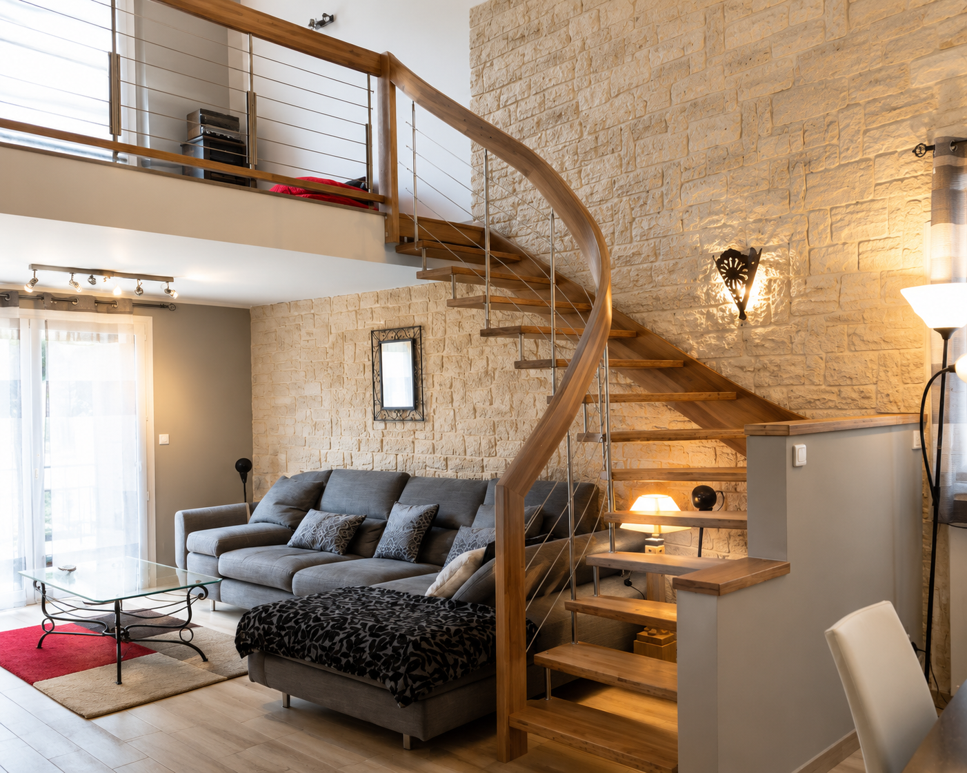 Salon moderne avec canapé gris, table basse en verre sur tapis coloré, escalier en bois courbé menant à une mezzanine, murs en pierre et éclairage chaleureux.