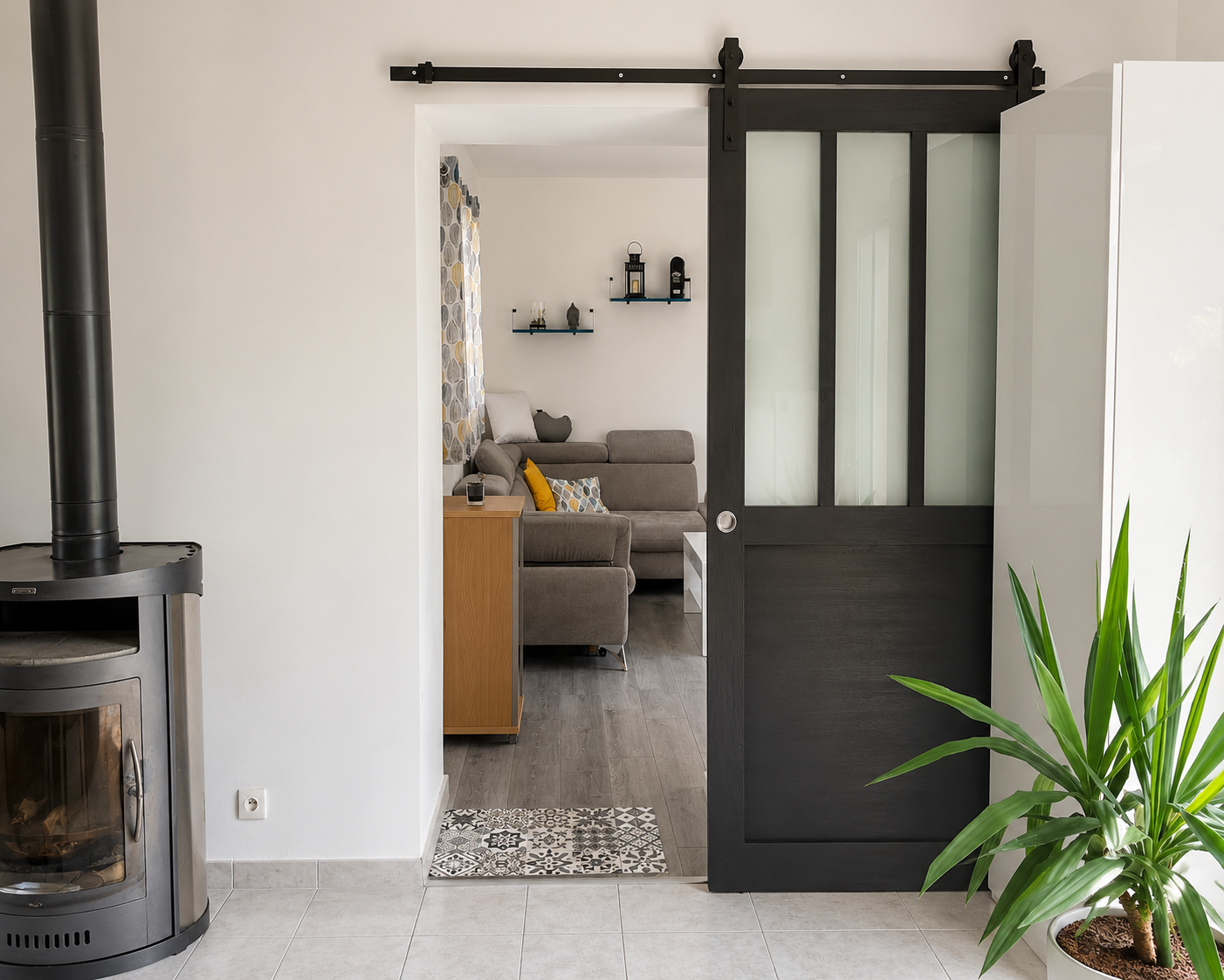 Intérieur moderne avec un poêle à bois noir à gauche, une porte coulissante noire avec panneaux translucides menant à un salon avec canapé gris, coussins colorés, meuble en bois et plante verte à droite.