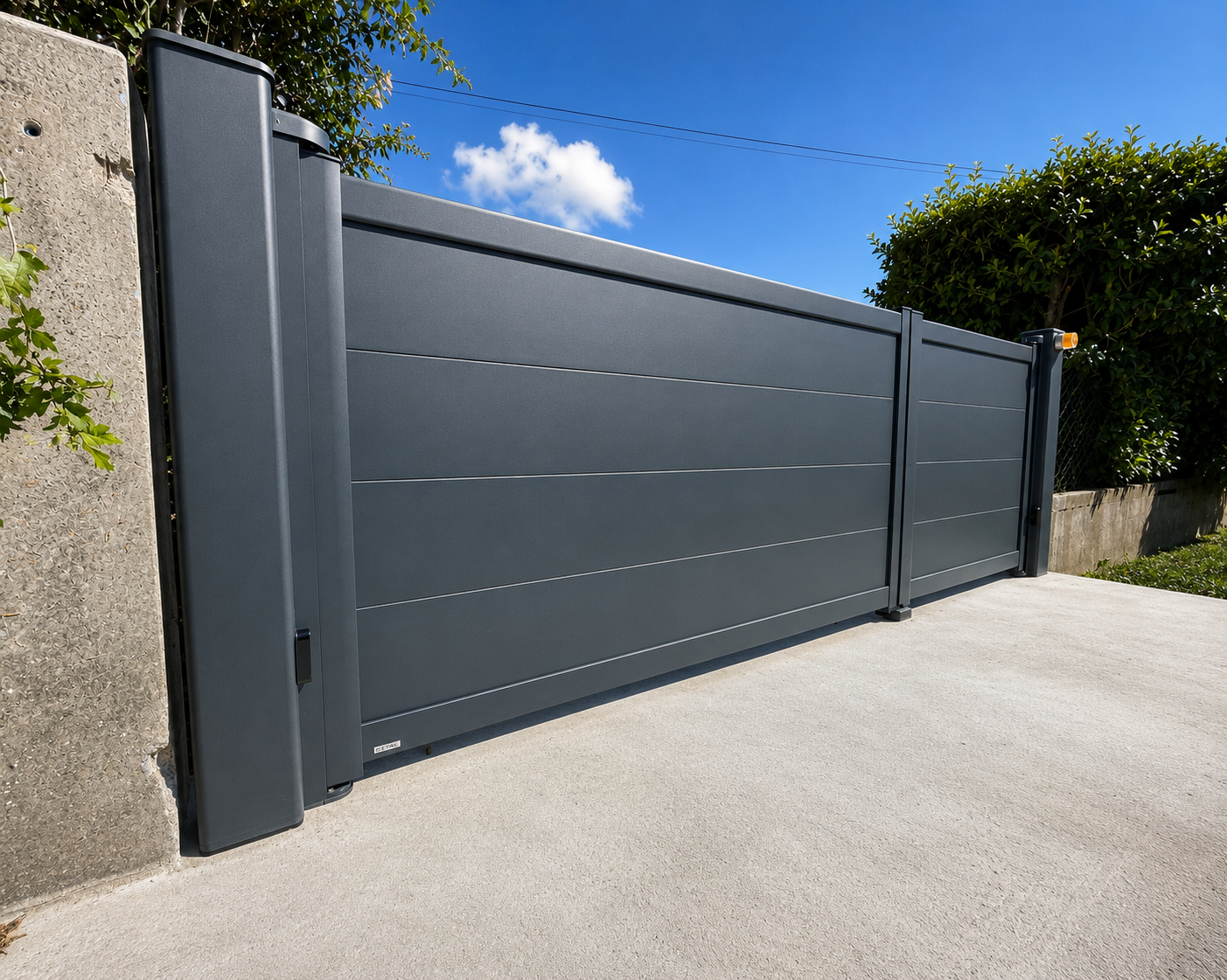 Portail motorisé gris foncé moderne installé sur une allée en béton sous un ciel bleu avec un buisson vert à droite.