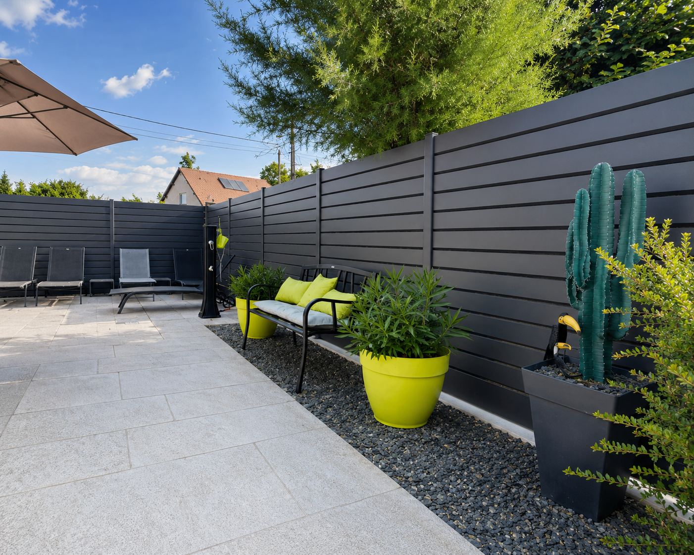 Terrasse extérieure avec un banc noir, deux grandes jardinières jaunes avec plantes, un cactus en pot carré noir, quatre chaises longues noires sous un grand parasol et une clôture grise en arrière-plan.