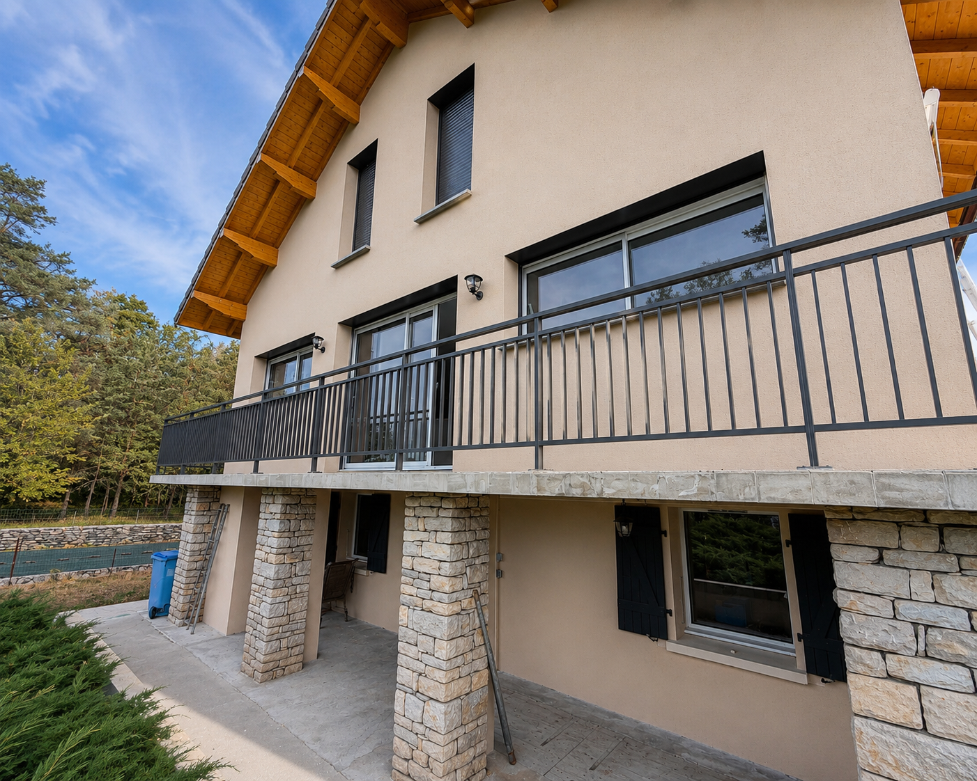Façade d’une maison moderne avec balcon en métal, murs beiges, piliers en pierres, fenêtres avec volets noirs et un toit en bois sous un ciel bleu clair.