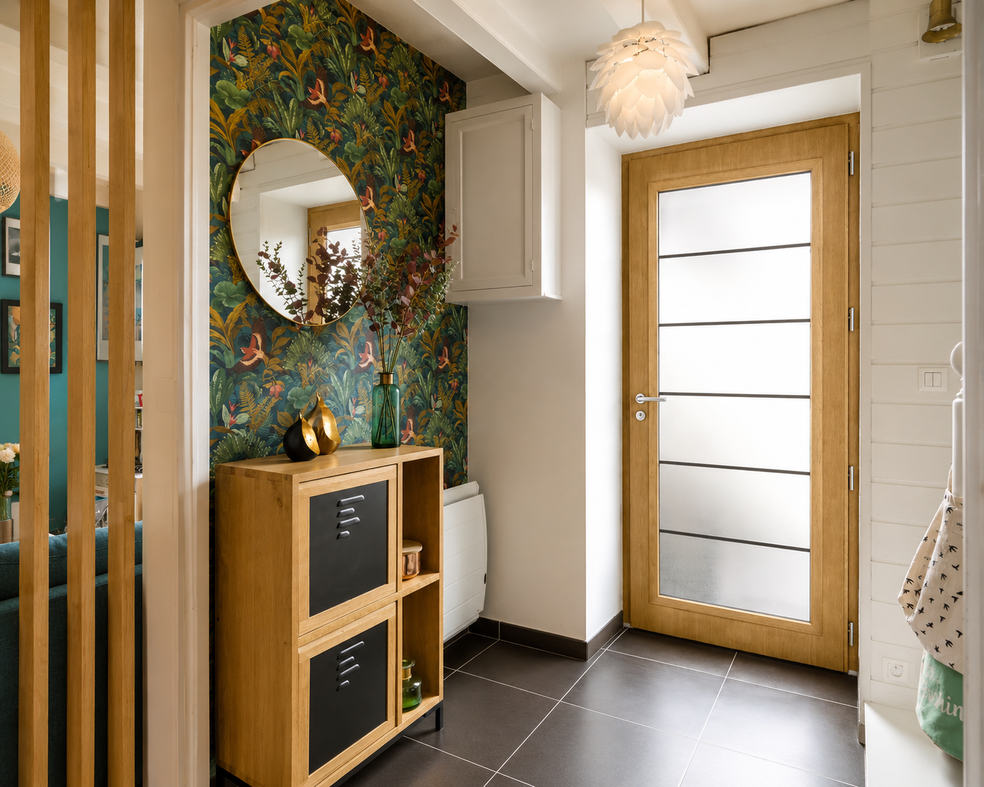 Entrée avec porte en bois clair à panneaux de verre dépoli, meuble de rangement en bois avec portes noires, mur décoré de papier peint végétal, miroir rond, vase vert avec branches, et lampe suspension blanche en forme de fleur.