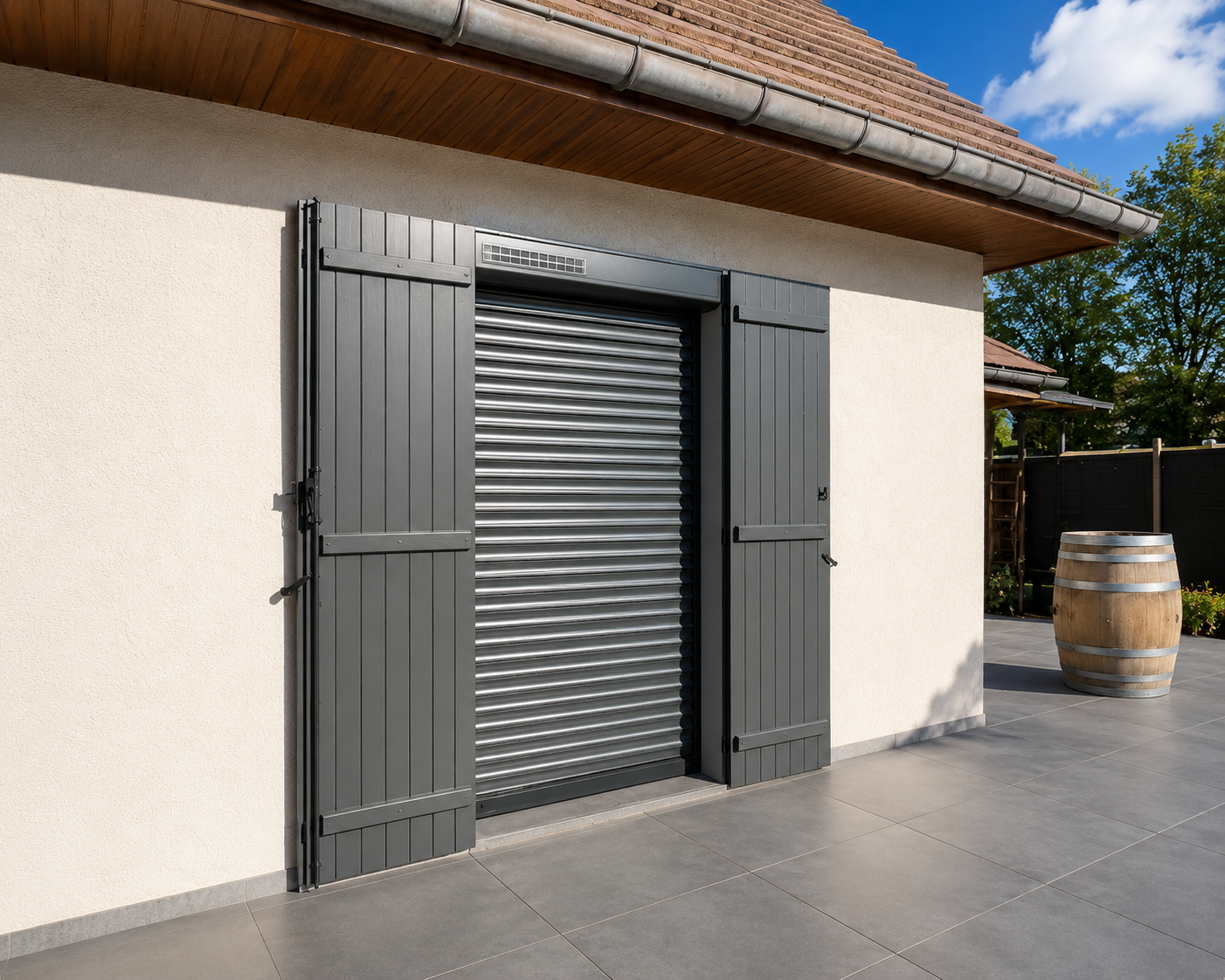 Volet roulant métallique gris installé sur une porte extérieure avec volets battants gris, au sol carrelé et un tonneau en bois à côté.