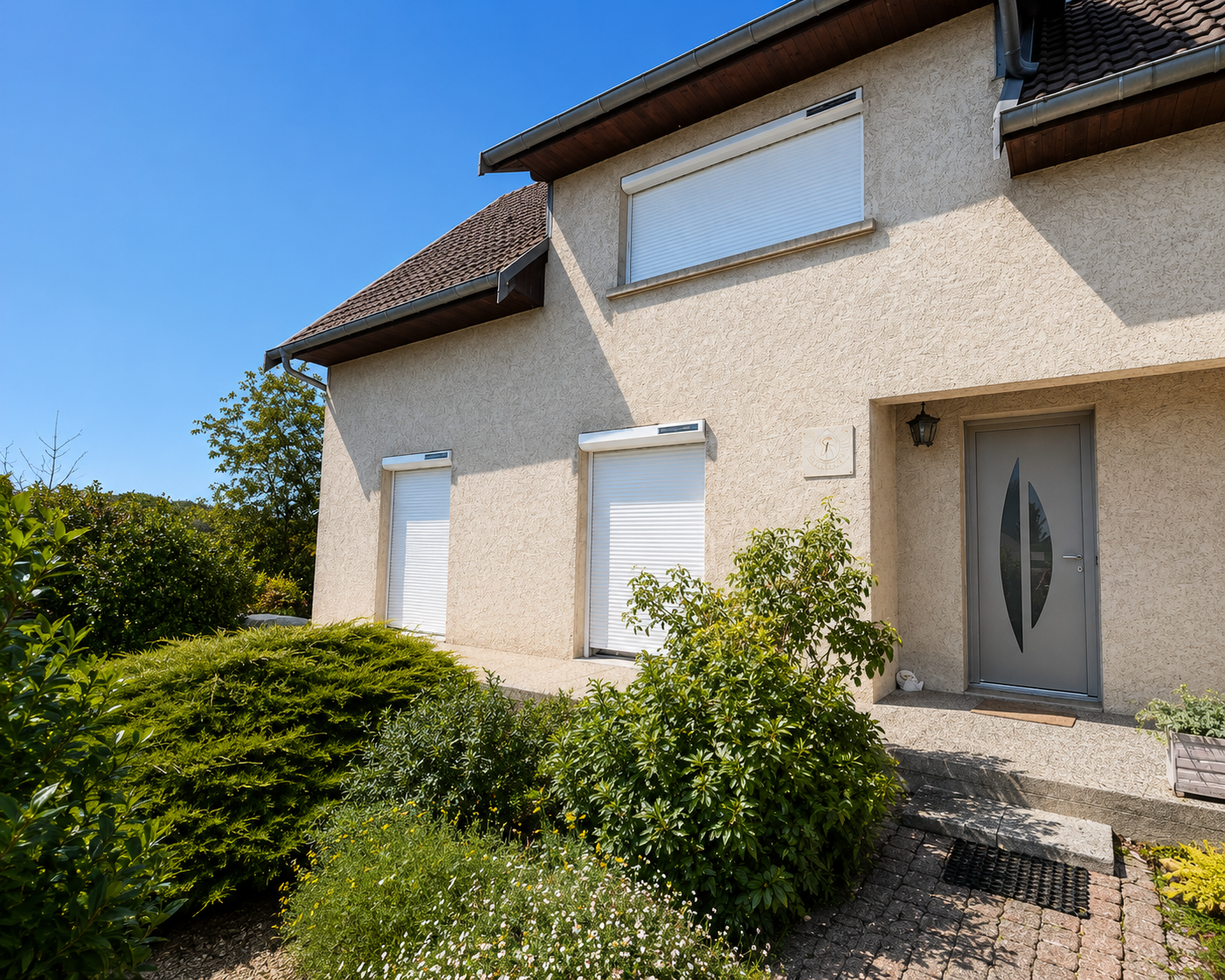 Façade d'une maison beige avec trois fenêtres à volets roulants blancs fermés, une porte grise moderne et un jardin avec des buissons et fleurs sous un ciel bleu clair.