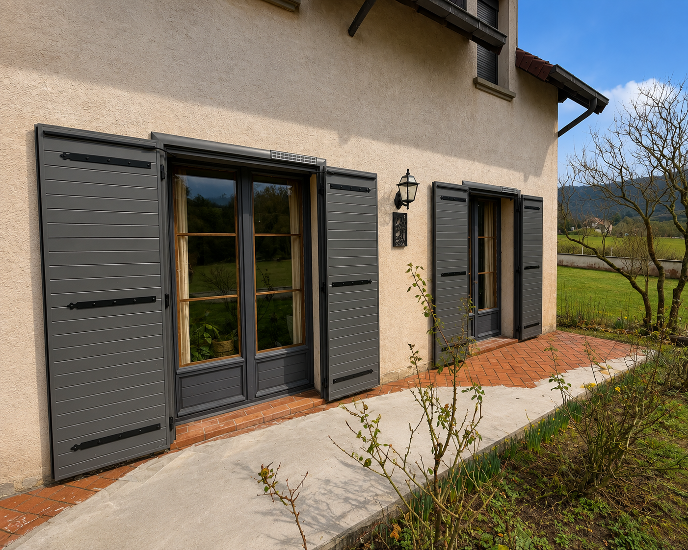 Façade extérieure d'une maison avec deux grandes portes-fenêtres en bois gris et volets ouverts, terrasse en briques et béton, arbustes devant et paysage de campagne en arrière-plan sous un ciel bleu.