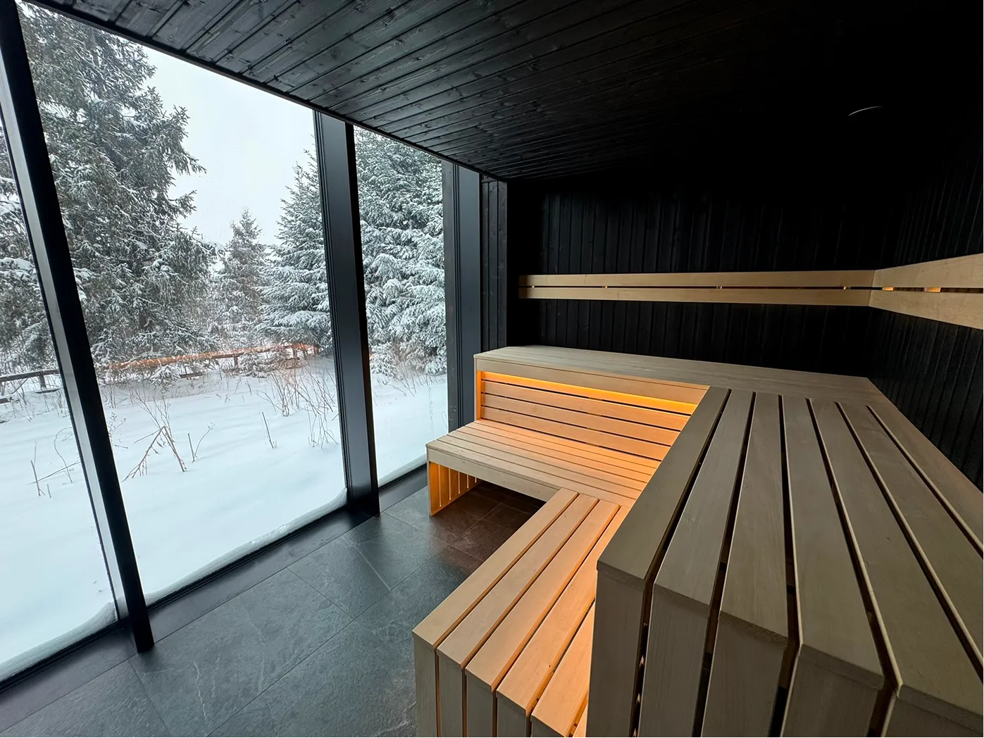 Wnętrze nowoczesnej suchej sauny z drewnianymi ławkami i dużym oknem, przez które widać zaśnieżone drzewa.