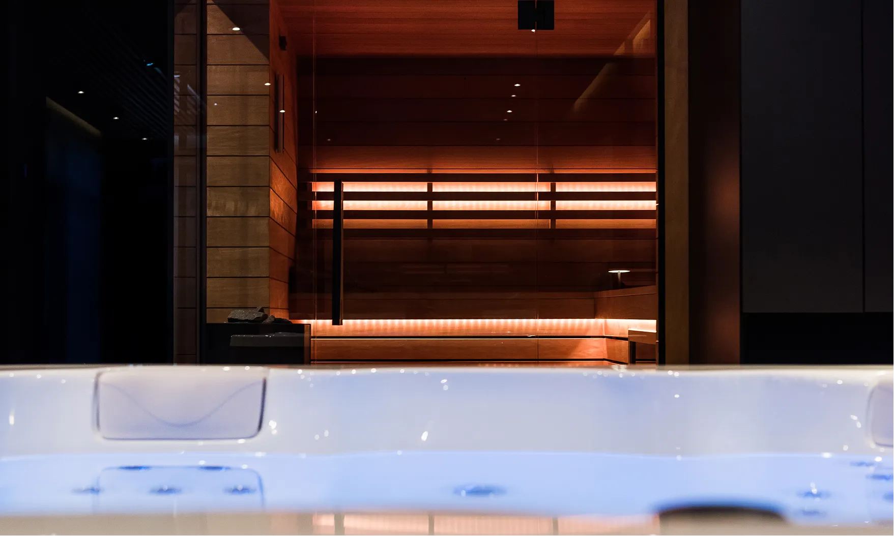 Nowoczesna sauna na podczerwień za przezroczystą szybą z oświetleniem LED i jacuzzi na pierwszym planie.