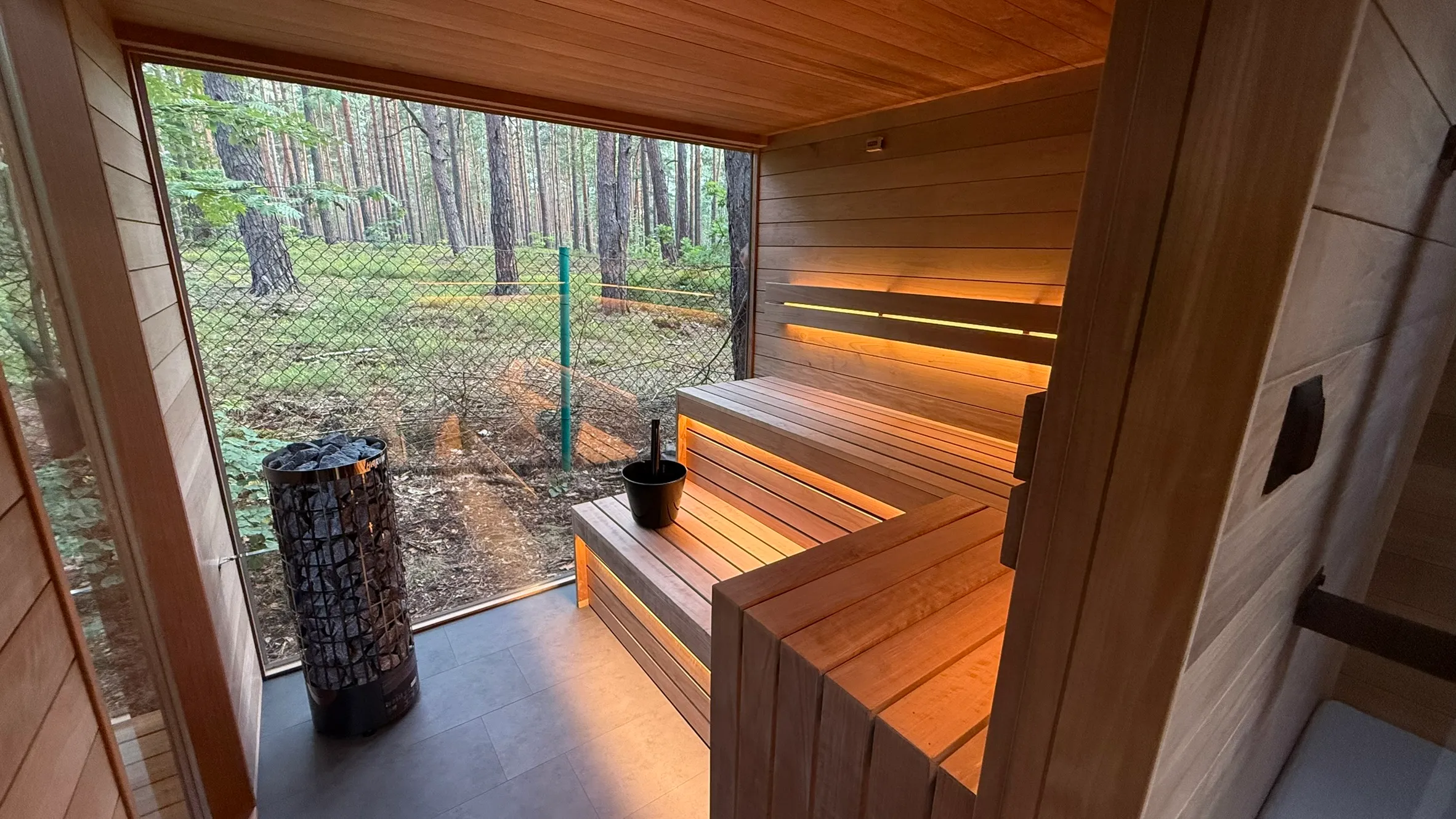 Nowoczesna sauna z drewnianymi ławkami i podświetleniem, widok na las przez szklaną ścianę.