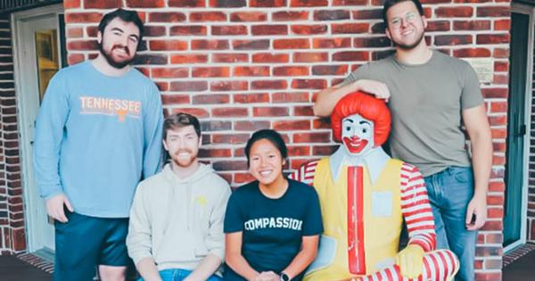 Four young adults pose for a photo next to a seated Ronald McDonald statue in front of a red brick wall. One person is standing on each end, and two are seated in the middle.
