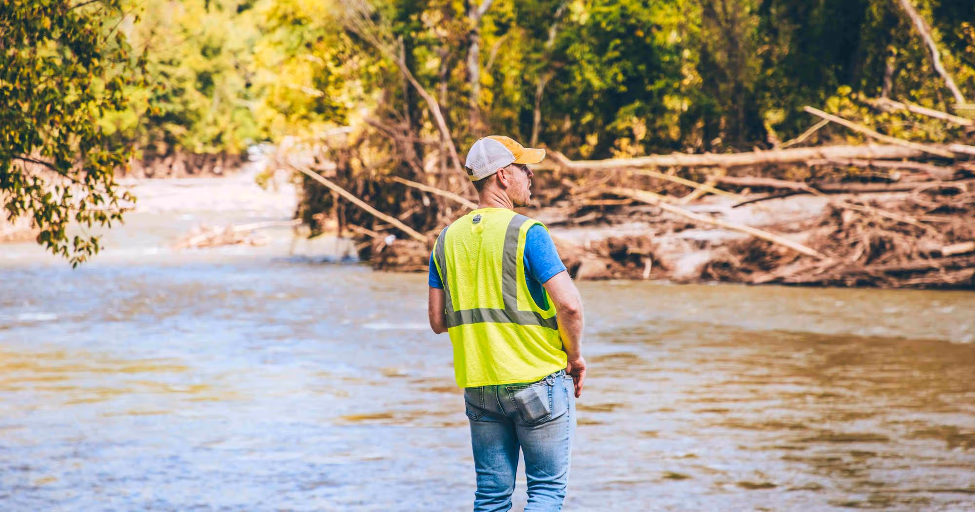 A man in a reflective vest stands on the bank of a river, surveying the scene after a flood.