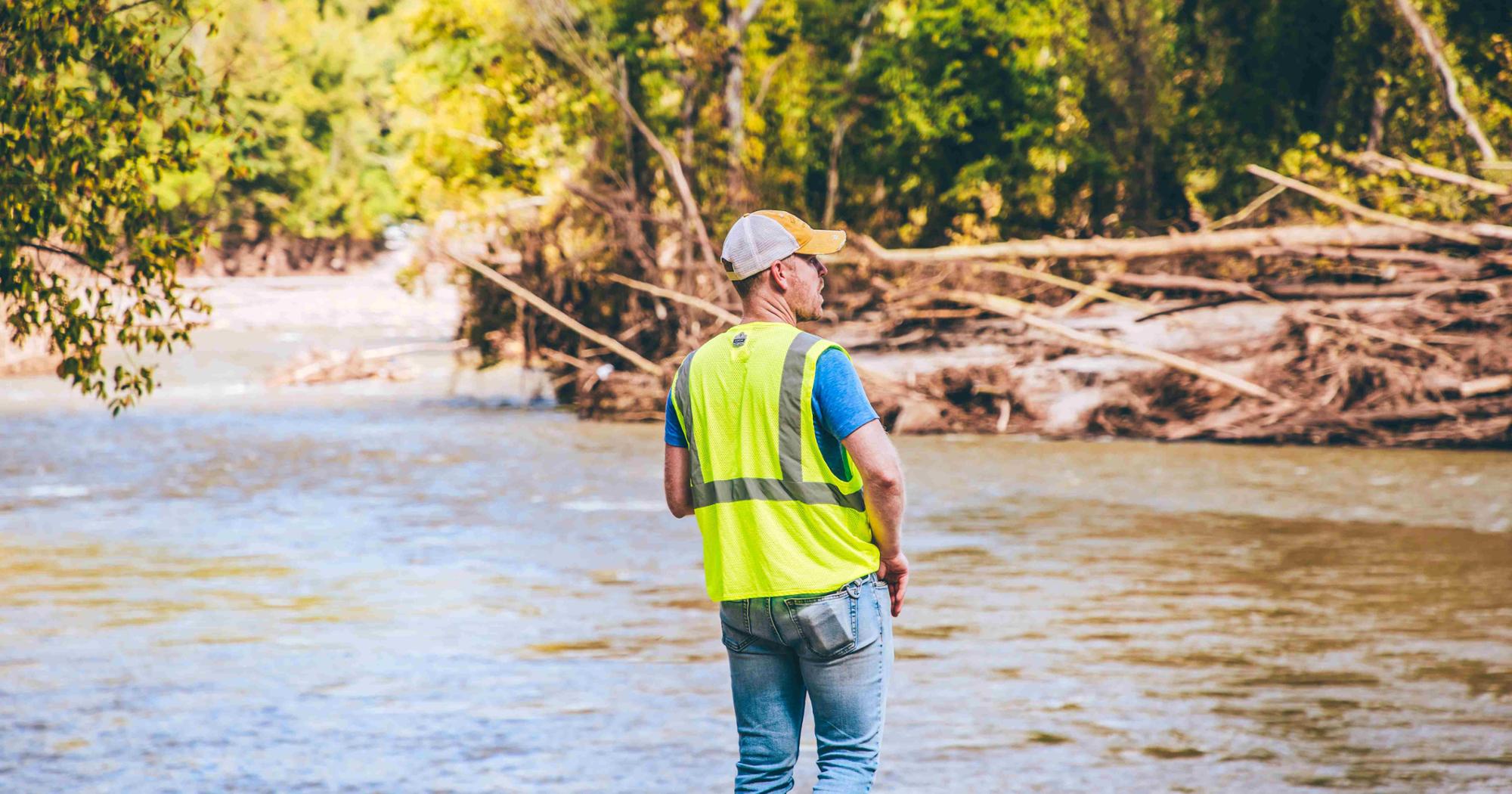 A man in a reflective vest stands on the bank of a river, surveying the scene after a flood.