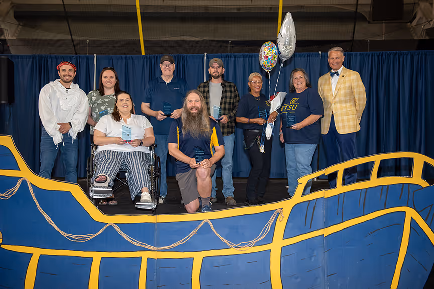 A group photo of ETSU’s 2025 Distinguished Staff Award winners and officials with a blue and gold pirate ship prop in the foreground reflective of the Staff Celebration theme, “Buccaneer Voyage.” Pictured (L-R) are ETSU Staff Senate President David Finney; award winners Morgan Kidd, Dr. Melody Blevins, Chuck Patton, John P. Lane, Casey Gwyn, Janice Kendrick and Jeri Paddock; and ETSU President Dr. Brian Noland. Not pictured: Jayson Hall.